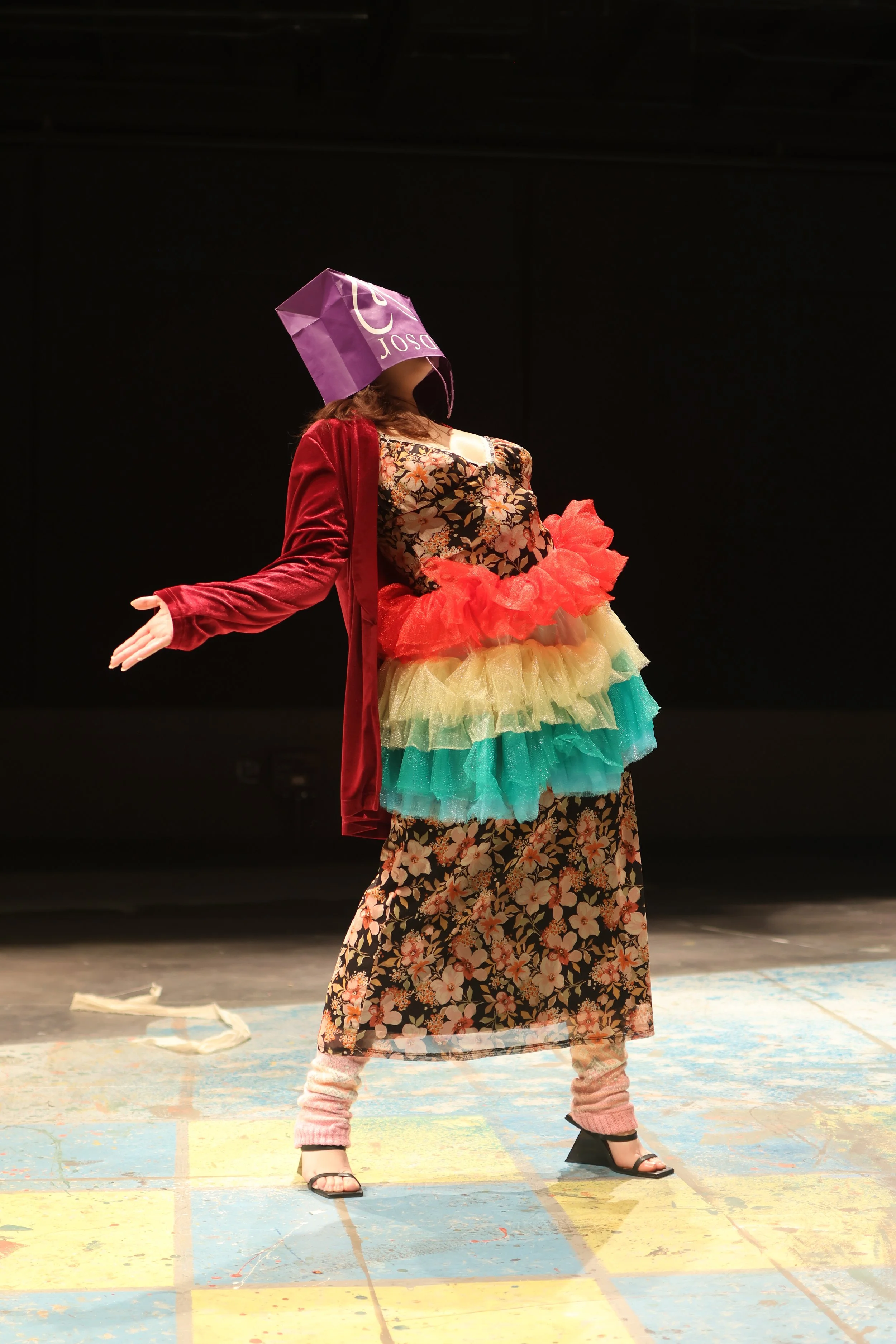 Young woman striking a silly pose on stage wearing a floral dress with rainbow-colored tutu, pink leg warmers, black heels, and a purple shopping bag on her head during a performance.