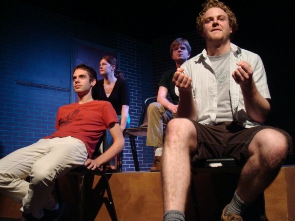 Four people on stage, with brick wall background, two sitting and two standing, in a theatrical performance.