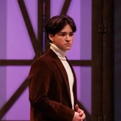 A young man on stage with wavy, dark hair, wearing a white turtleneck and brown blazer, standing in front of a purple background with wooden beams.