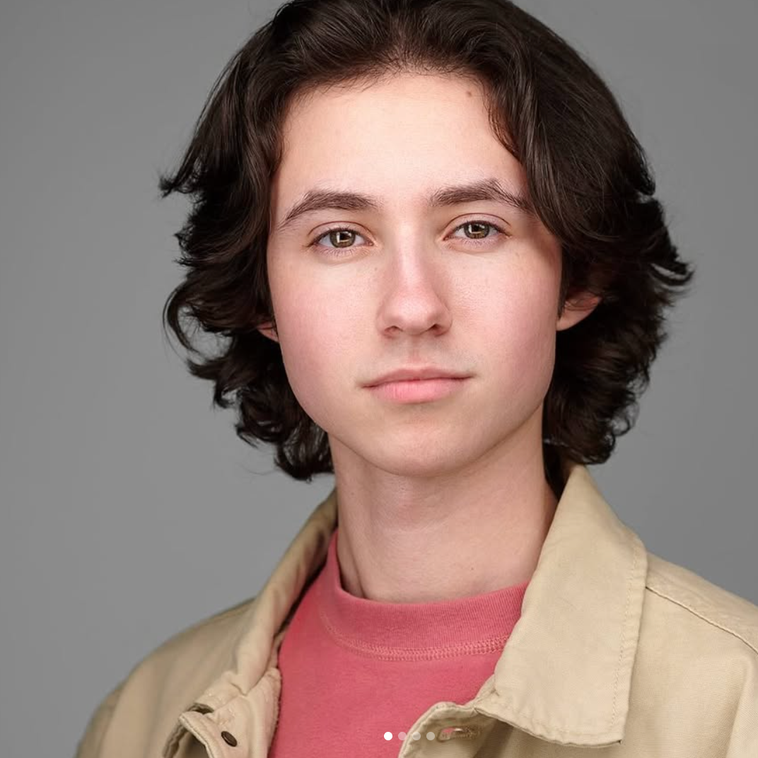 Headshot of a young person with medium-length wavy brown hair, wearing a beige jacket over a pink shirt, against a gray background.