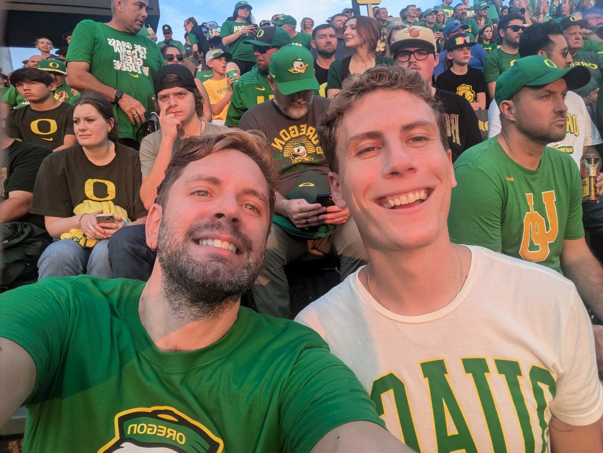 Crowd of sports fans dressed in green and black Oregon Ducks attire at a stadium, some smiling and engaging with each other.