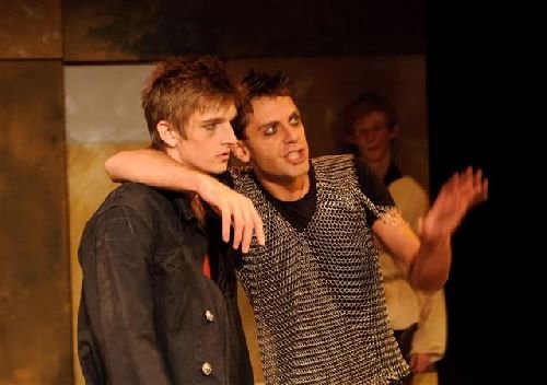 Two young men, one with brown hair wearing a dark jacket and the other with black hair wearing a patterned shirt, are sitting close together, with the man in the patterned shirt apparently speaking or explaining something. A person in the background looks on, smiling.
