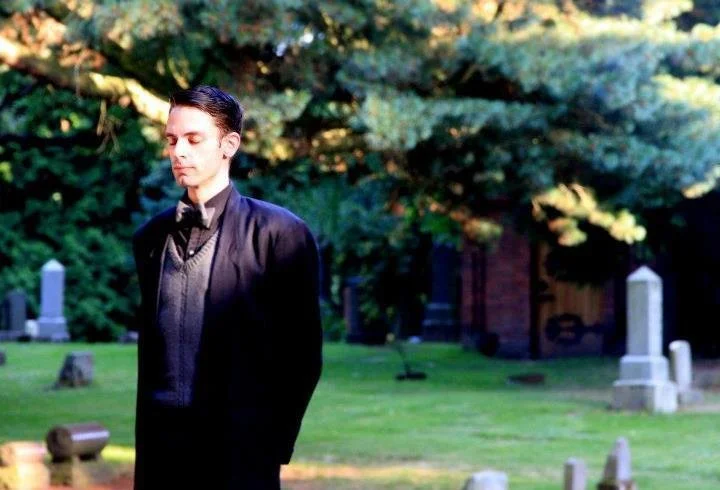 A man in a black suit with a bow tie standing in a graveyard with tombstones and trees.