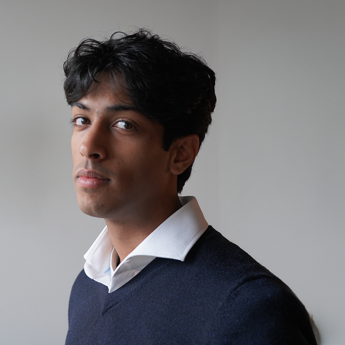 Close-up of a young man with dark wavy hair, wearing a white collared shirt and a navy sweater, standing against a plain light gray background.