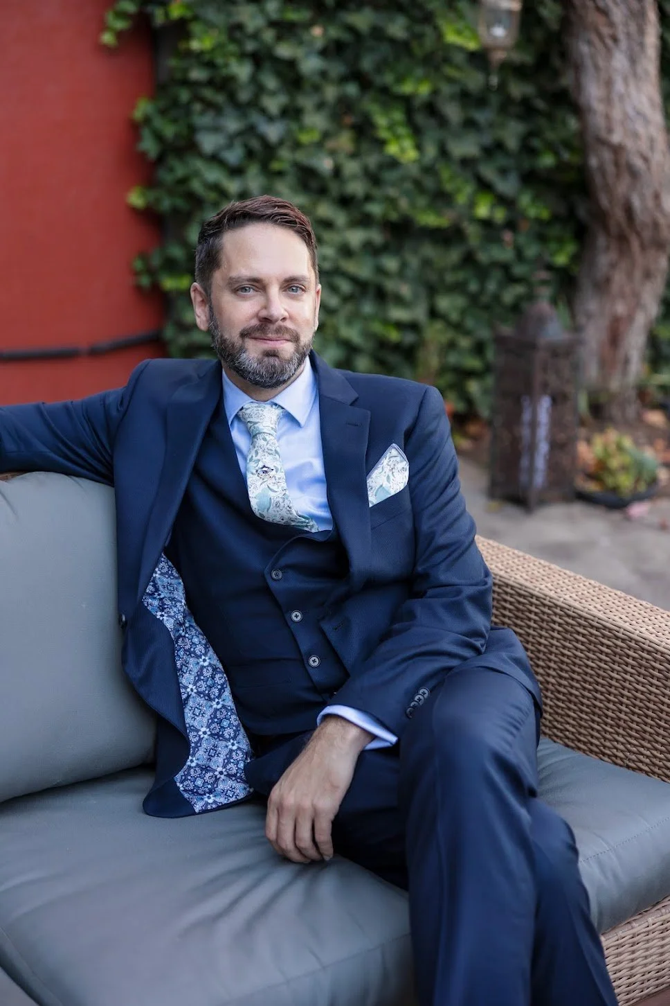 A man in a navy blue suit sitting on an outdoor wicker sofa with greenery and trees in the background.