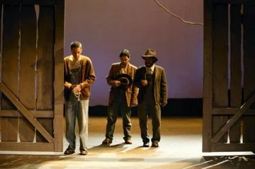 Three men standing on a stage in a theater, with doors on either side, one holding a drink, another holding a book, and the third wearing a hat, possibly rehearsing or performing.