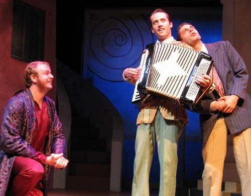 Three men performing on stage, one playing an accordion and two singing or acting, with a blue background and interior setting.