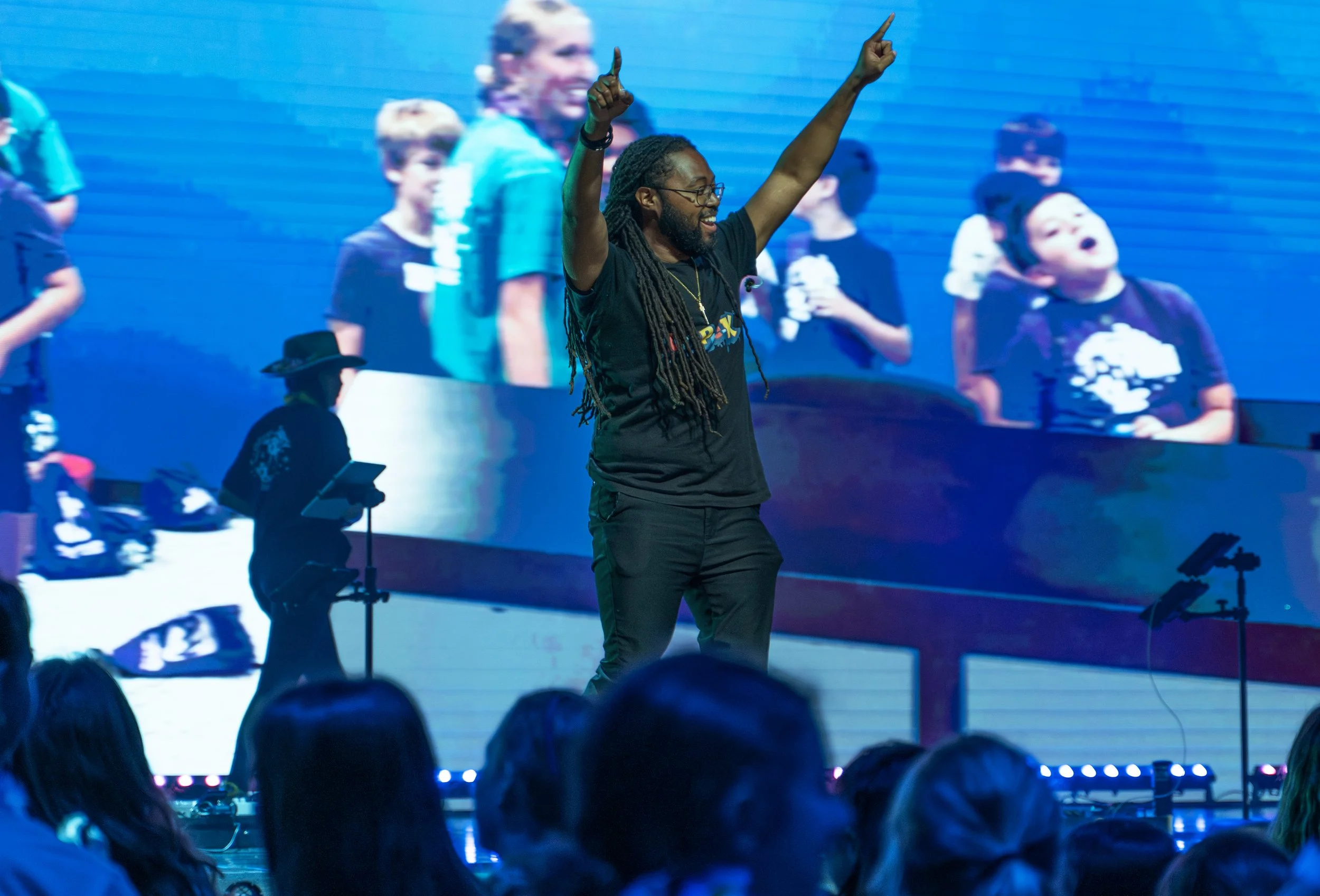 A man with dreadlocks and glasses is smiling and raising both arms with index fingers pointing upward on stage, with a large screen behind him displaying a blurred image of children and adults.
