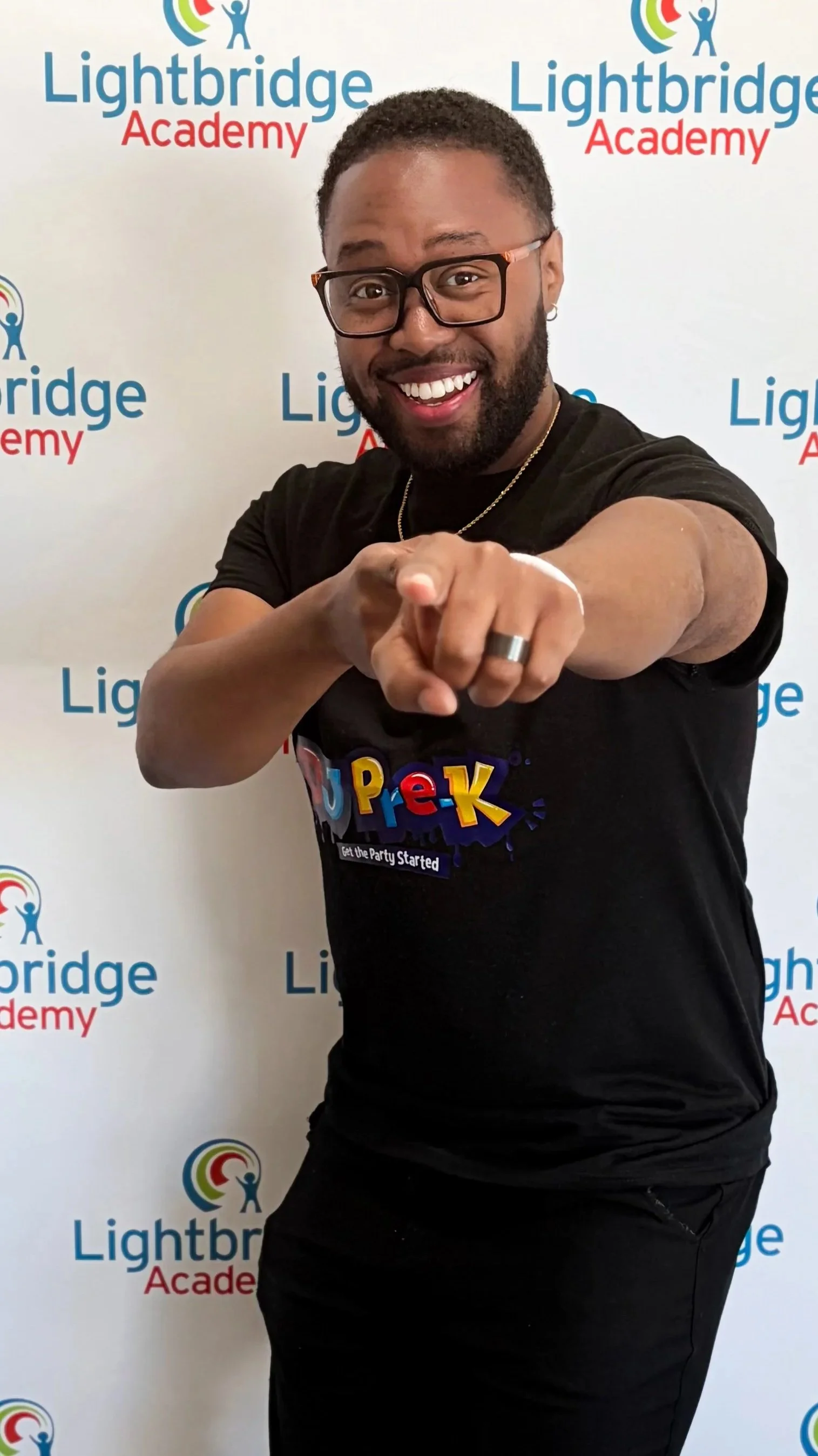 DJ Pre-K smiling and pointing at the camera in front of a Lightbridge Academy backdrop.