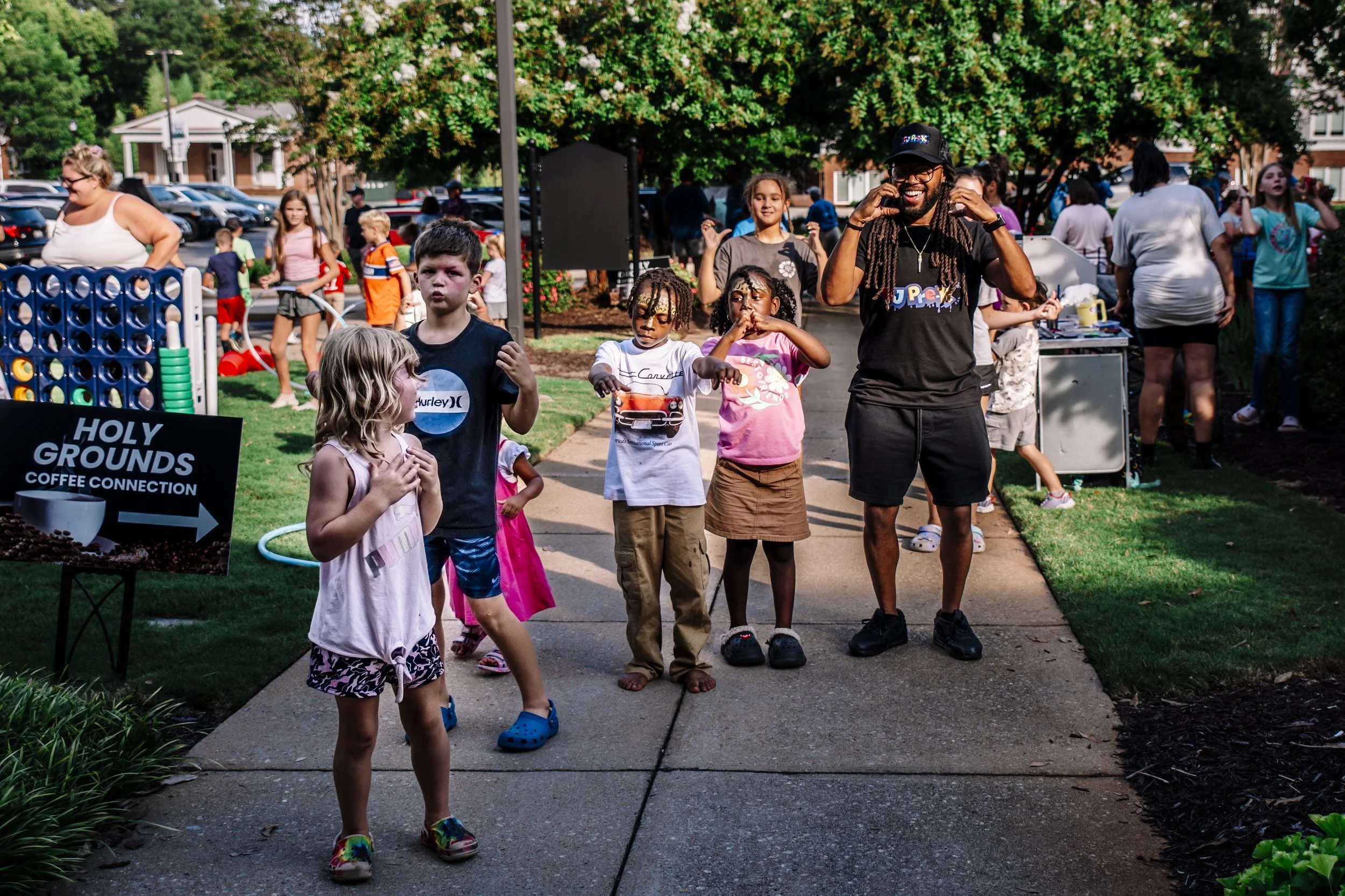 A diverse group of children and adults at an outdoor community event, with children standing in a line on a sidewalk, some talking and others observing, and adults in the background near booths and tables, surrounded by trees and parked cars.