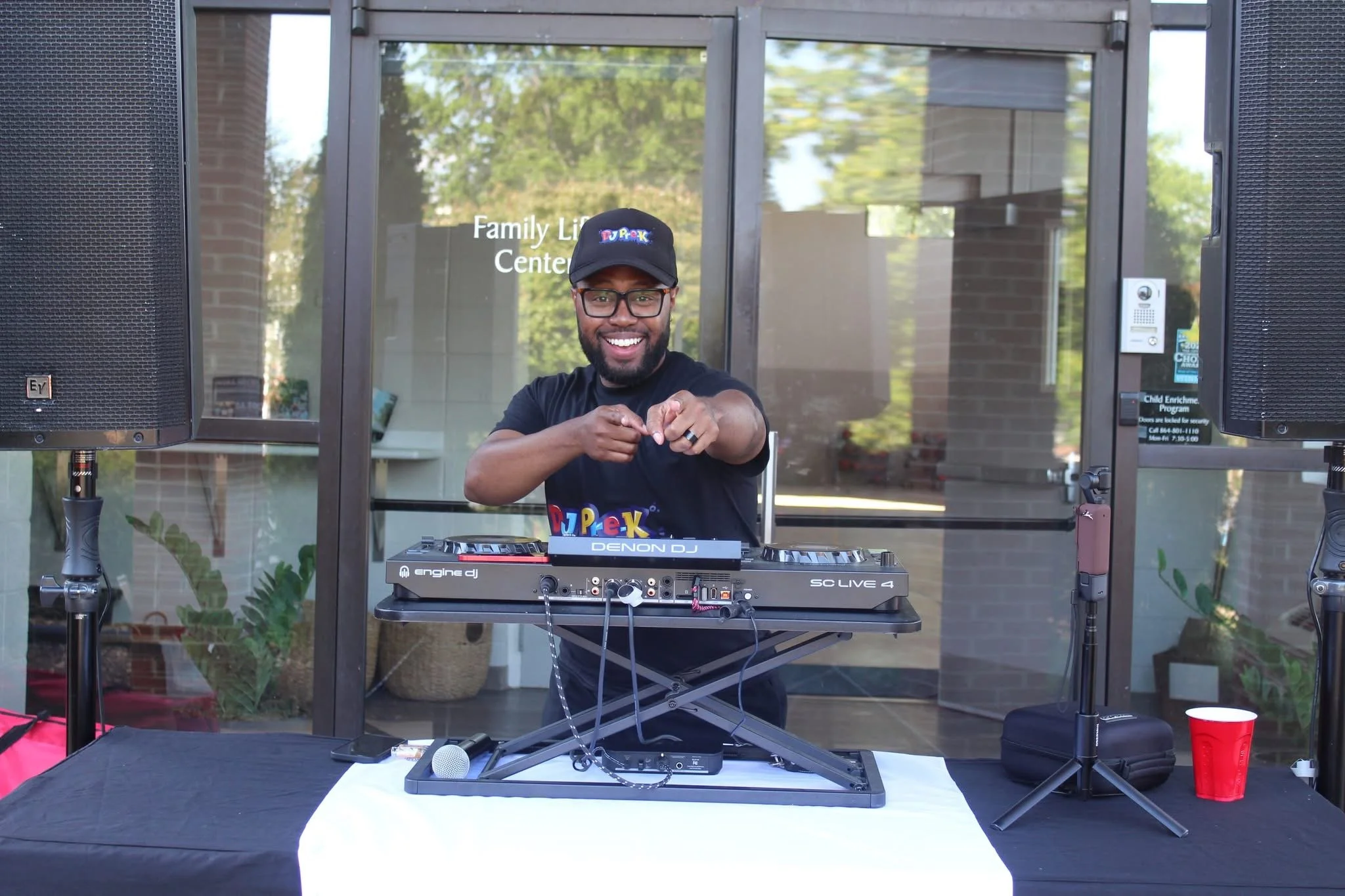 DJ Pre-K , smiling and pointing, standing behind a DJ setup with speakers on either side, in front of a building entrance with glass doors, trees, and a sign that reads 'Family Life Center'.