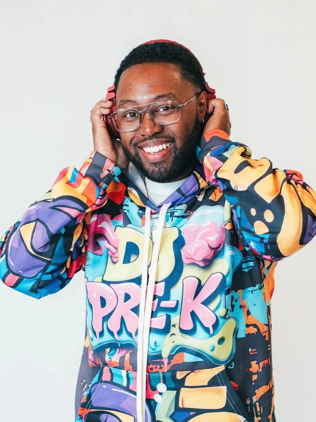 DJ Pre-K smiling wearing a colorful graffiti-style hoodie with the words 'DJ PRE-K' on it, holding red headphones to his ears.