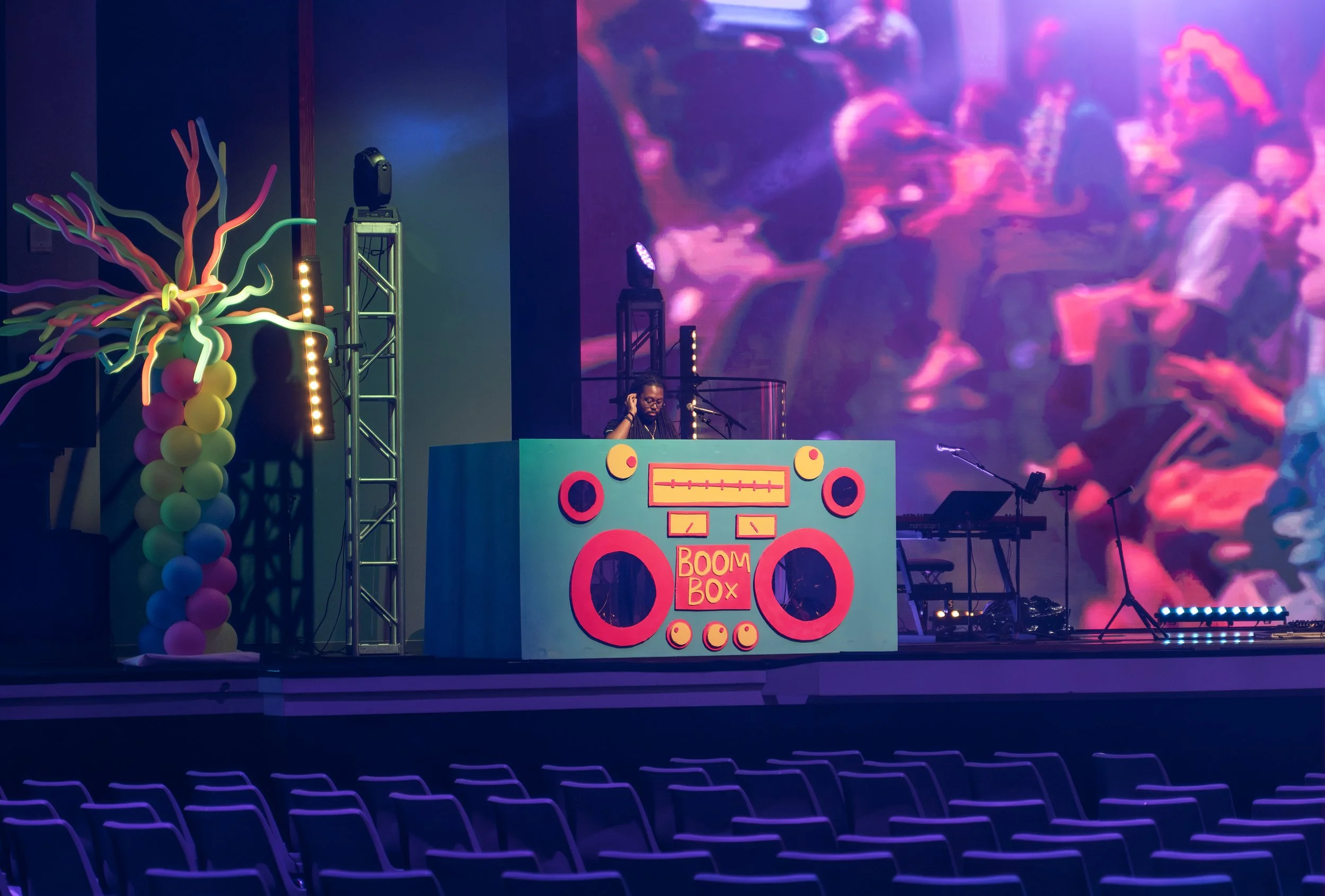 Empty auditorium with a stage featuring a colorful DJ booth labeled 'BOOM BOX,' a performer with long hair behind it, neon balloon decorations, and a large digital screen displaying abstract visuals.