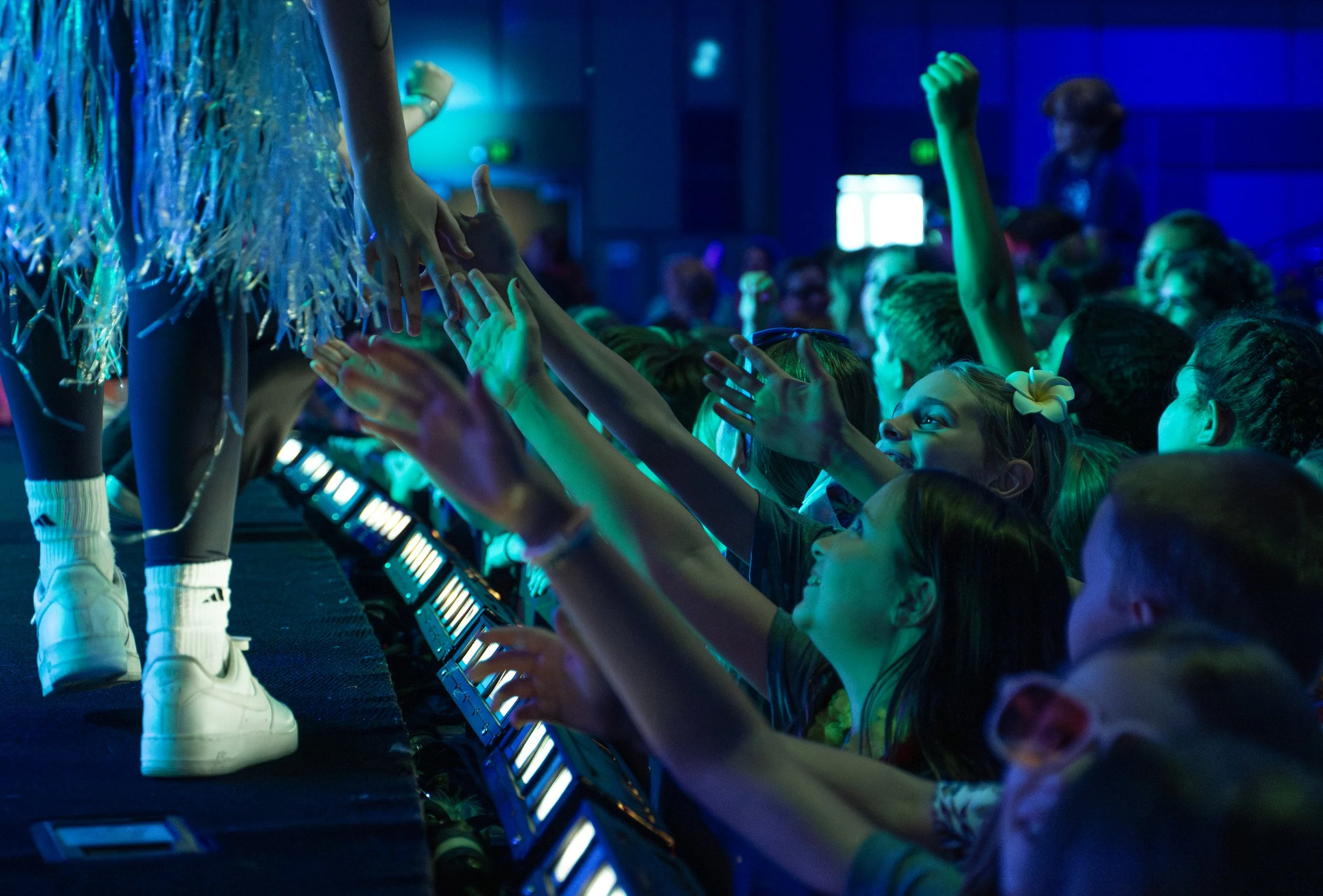 A group of children reaching out their hands towards performers on stage in a dark, colorful environment, possibly at a concert or event.