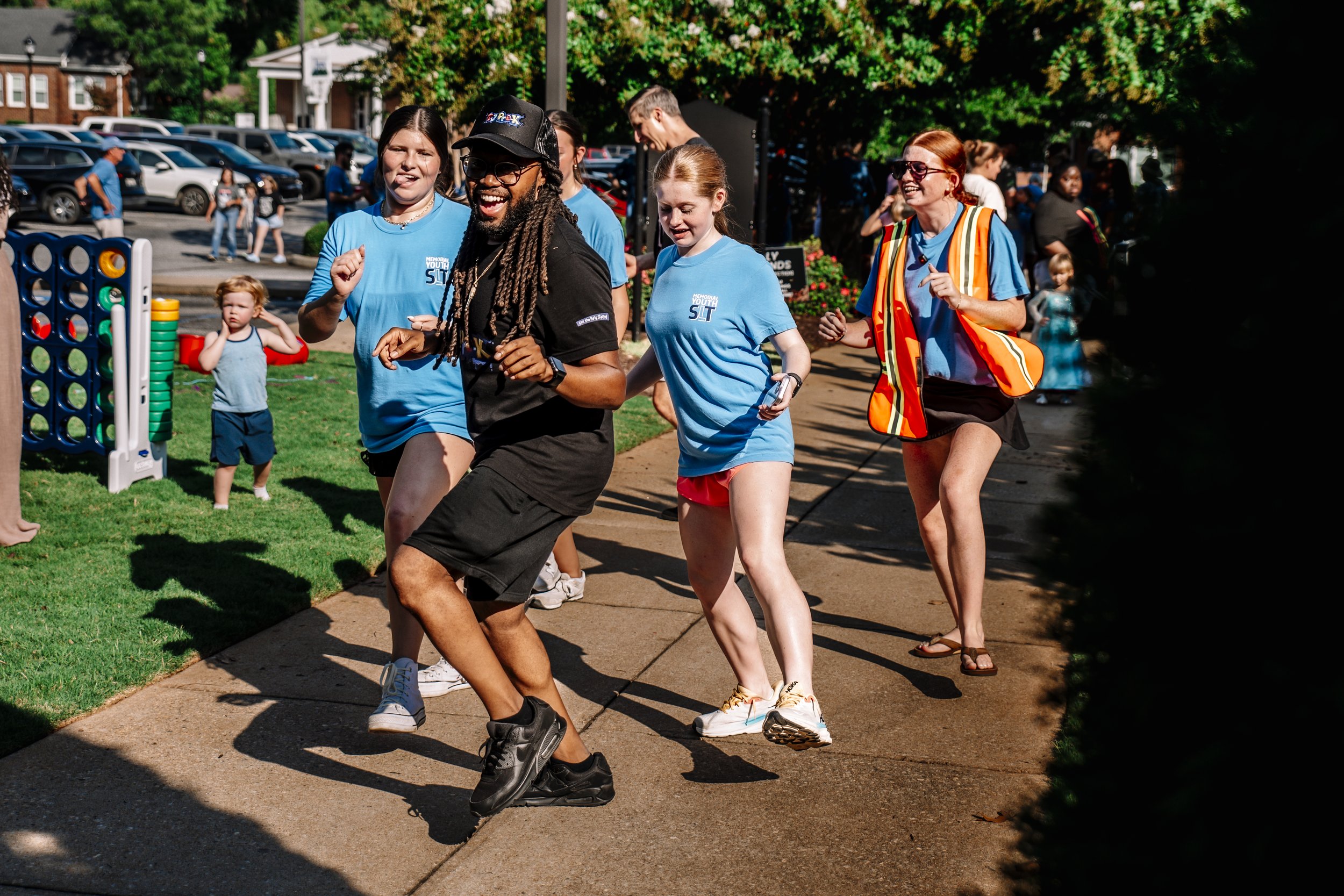 People participating in a running event outdoors on a sunny day, with cars parked and trees in the background.
