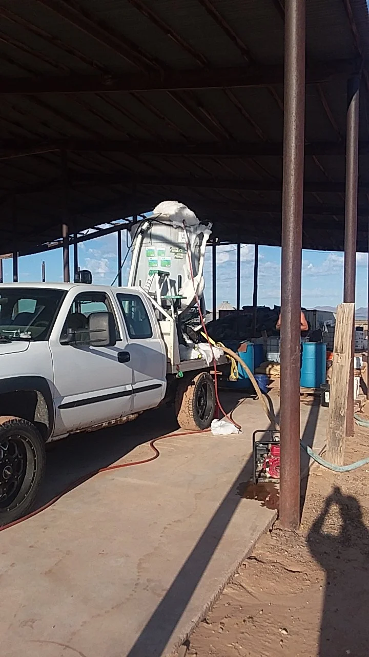 A white truck with equipment on the back under a shaded structure. The back of the truck is open with a foam object on top, and hoses are connected to the equipment, with some on the ground. There are blue barrels and debris nearby.