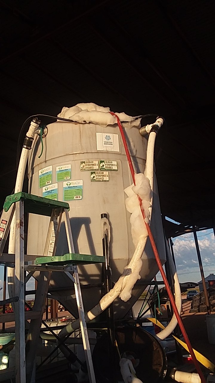 Large oval-shaped tank with multiple labeled stickers, foam insulation, and connected piping, set outdoors with a ladder leaning against it and a partly cloudy sky in the background.