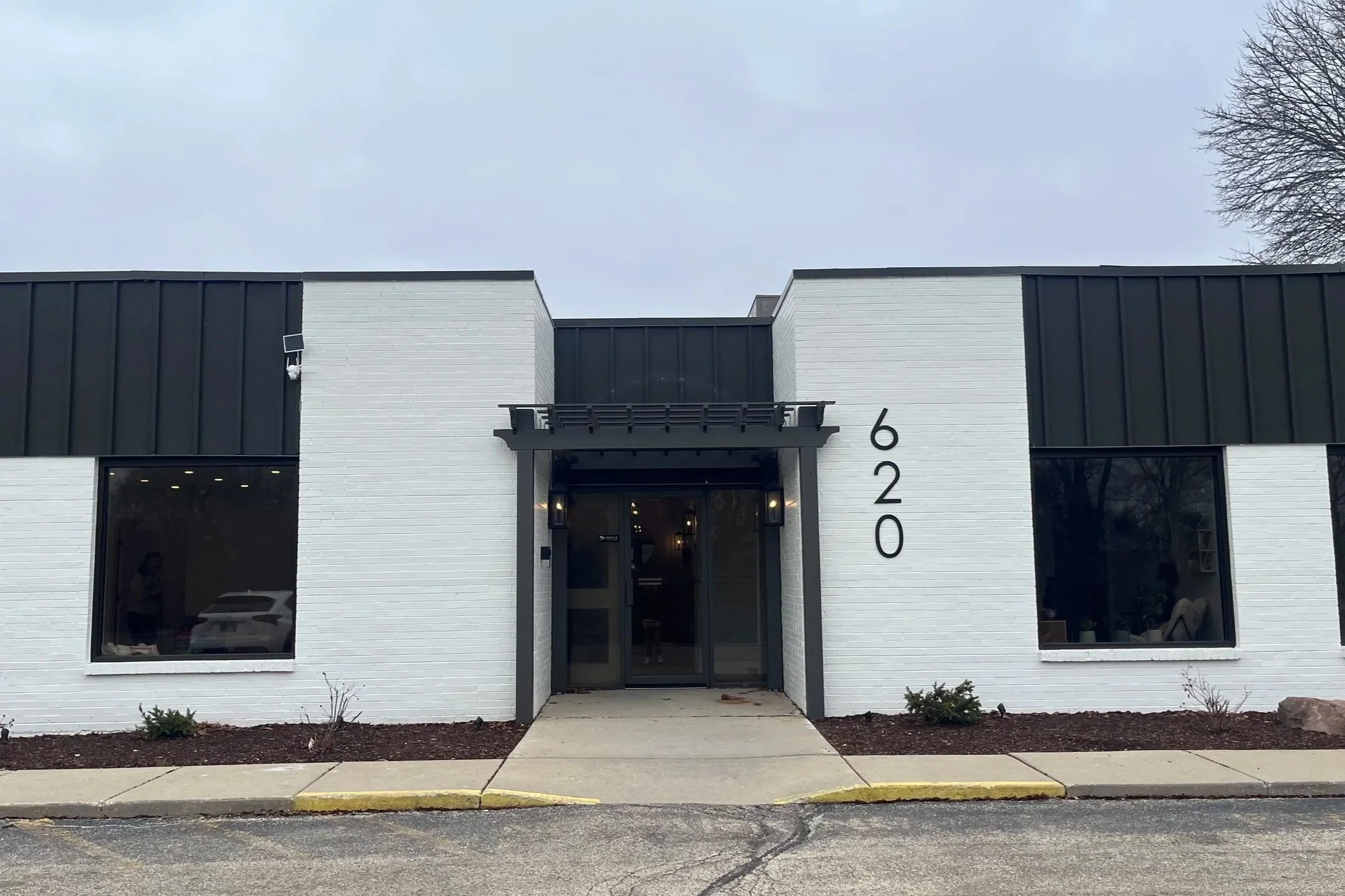 A modern building with white brick walls and black accents, including large windows and a central entrance door, with the street address 620 displayed vertically on the right side of the building. The sidewalk and street are visible in the foreground, with bare trees and a cloudy sky in the background.