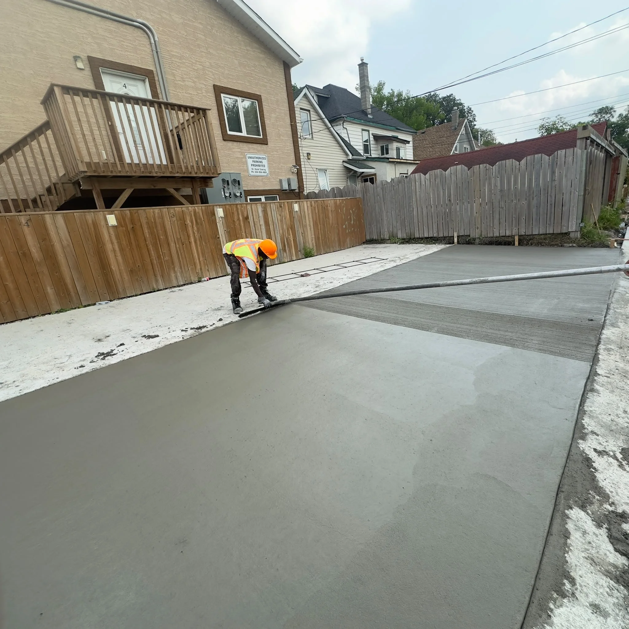 A worker in a safety vest and helmet smoothing fresh concrete in a driveway with a wooden fence and houses in the background.