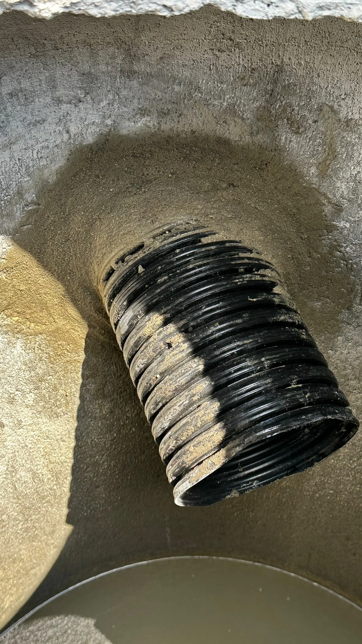 A close-up of a large black corrugated pipe with dirt and mud inside, connected to a concrete surface, with a small amount of water at the bottom.