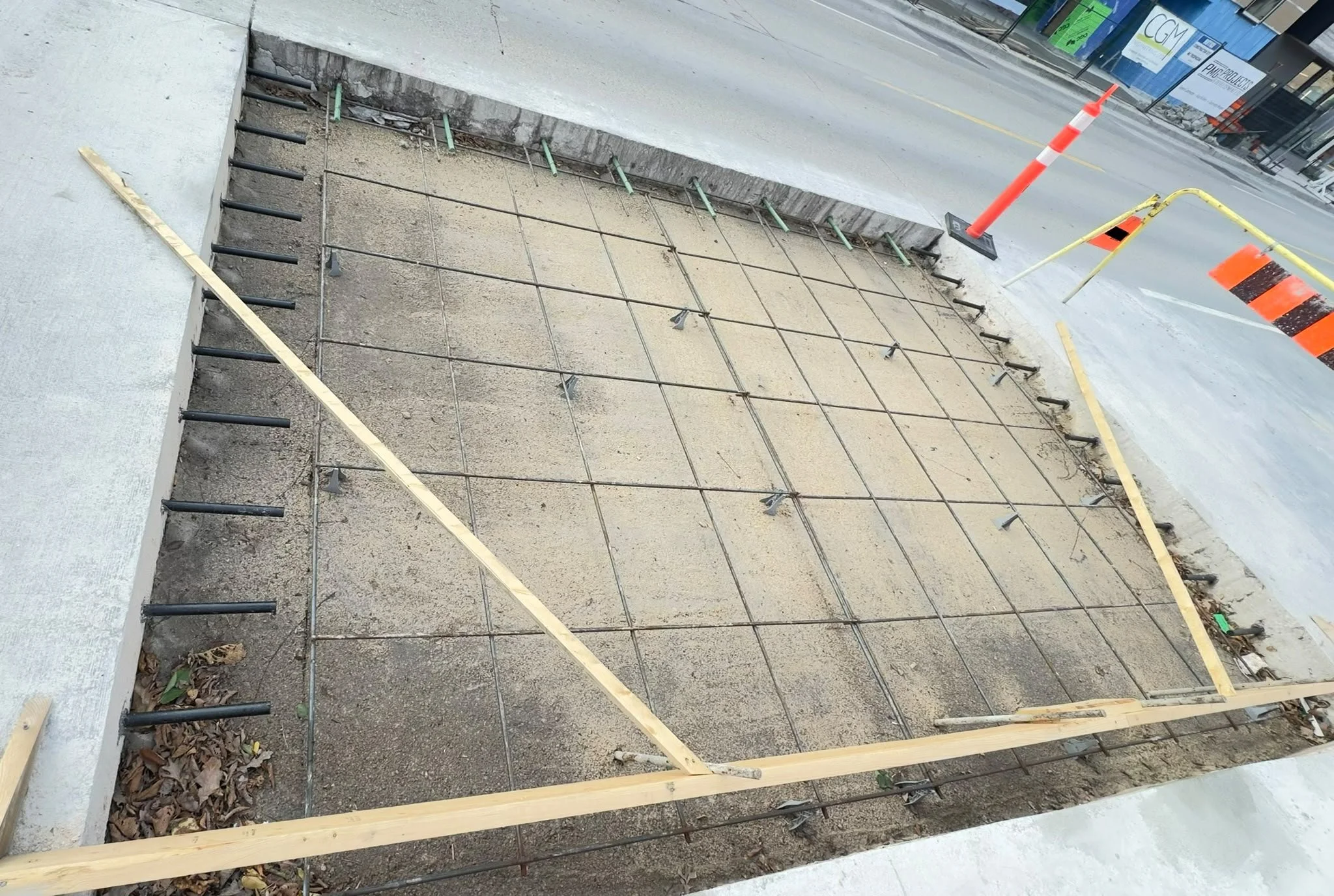 Construction site showing a sidewalk section with rebar and wooden forms, surrounded by orange fencing and traffic cones.