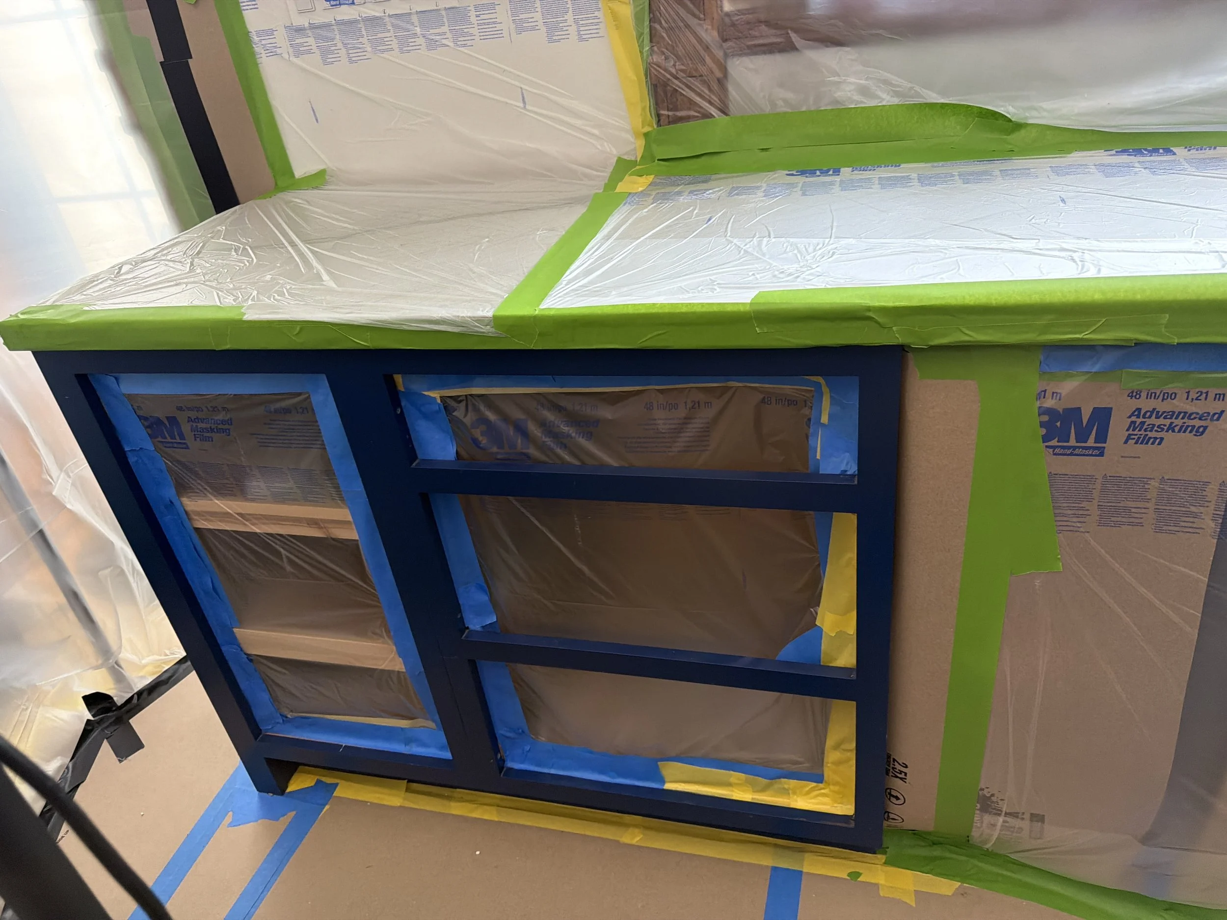 A blue wooden frame of furniture or a display case wrapped in clear plastic and green painter's tape, with cardboard boxes labeled '3M Advanced Masking Film' in the background, indicating ongoing construction or renovation work.