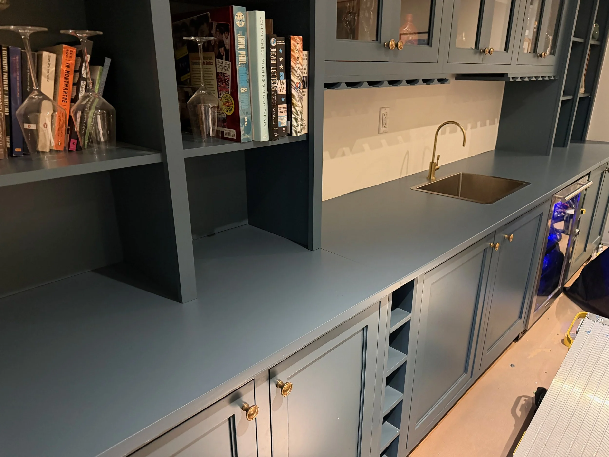 A partially assembled blue kitchen cabinet with open shelves, gold knobs, and a built-in wine rack. Several books and upside-down wine glasses are on the top shelf.