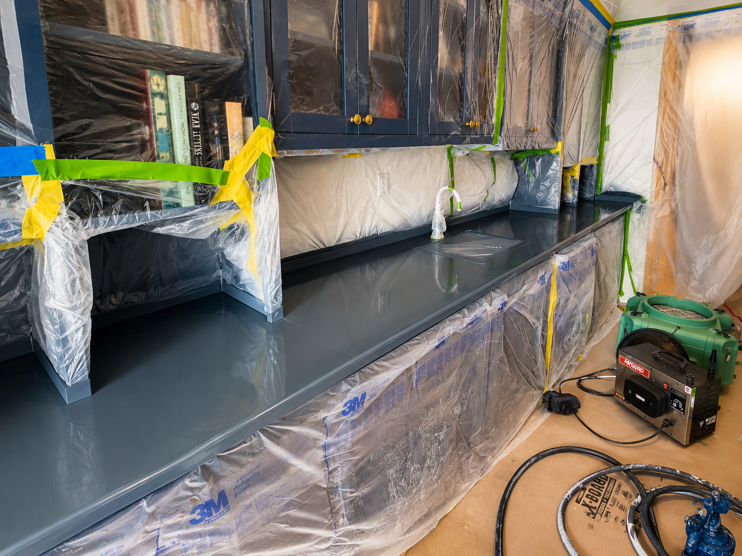 Kitchen renovation in progress with cabinets and countertops covered in plastic sheeting for protection, and construction tools and equipment on the floor.