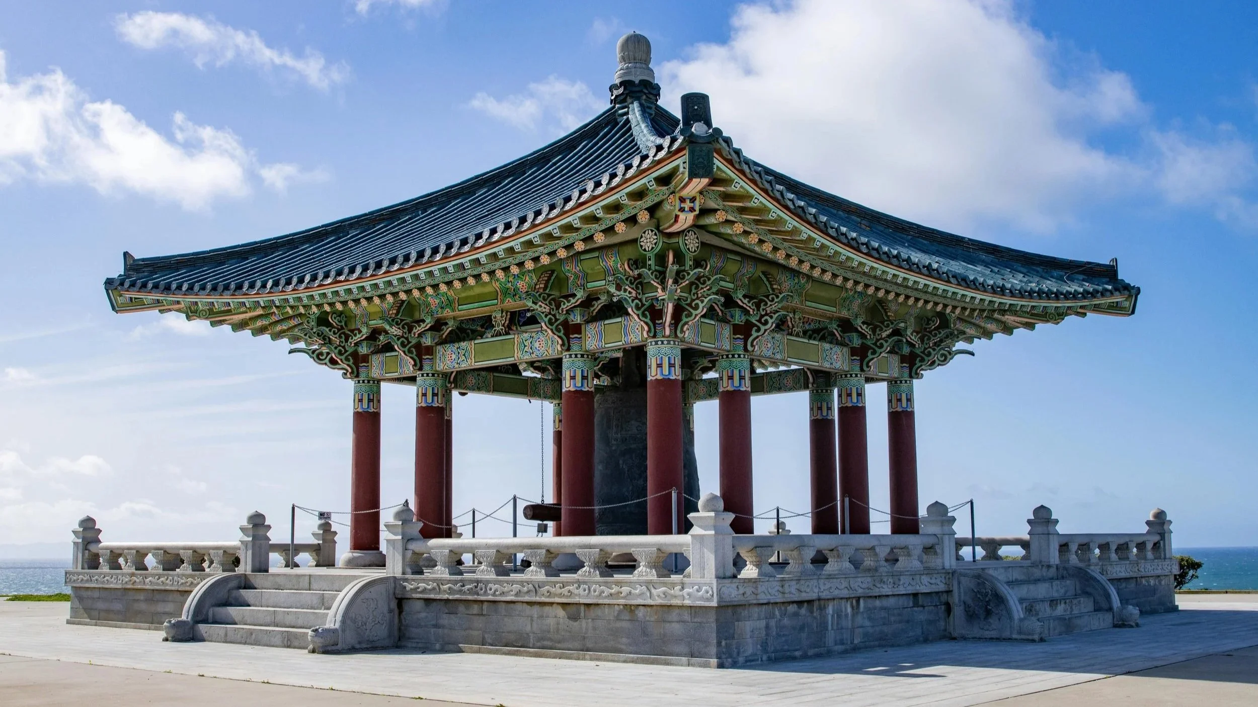 A traditional Asian-style pavilion with intricate, colorful designs on the roof and supporting pillars, situated outdoors with a view of the ocean under a partly cloudy sky.