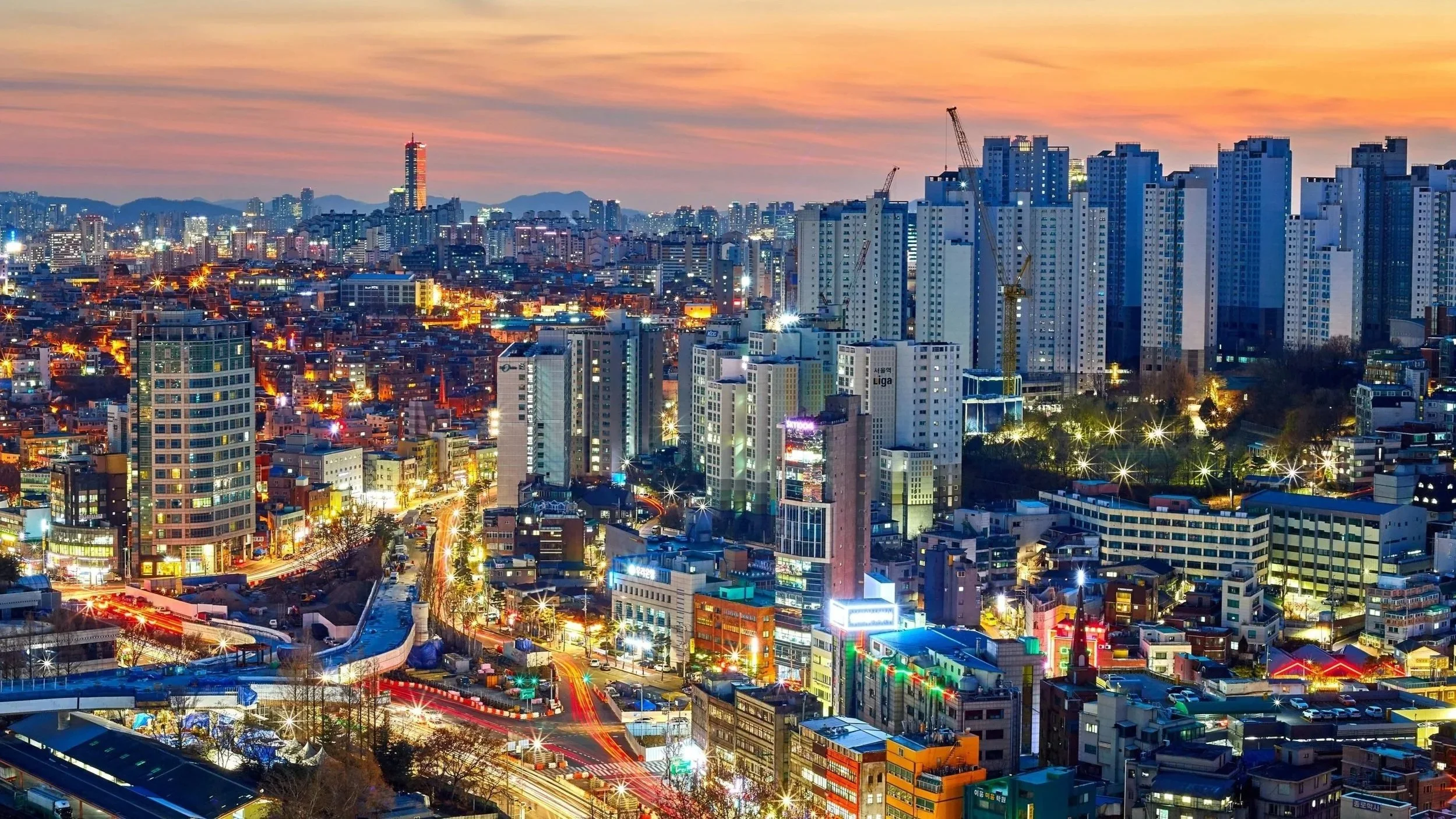 A cityscape at dusk with tall buildings, construction cranes, and colorful lights illuminating the streets below.