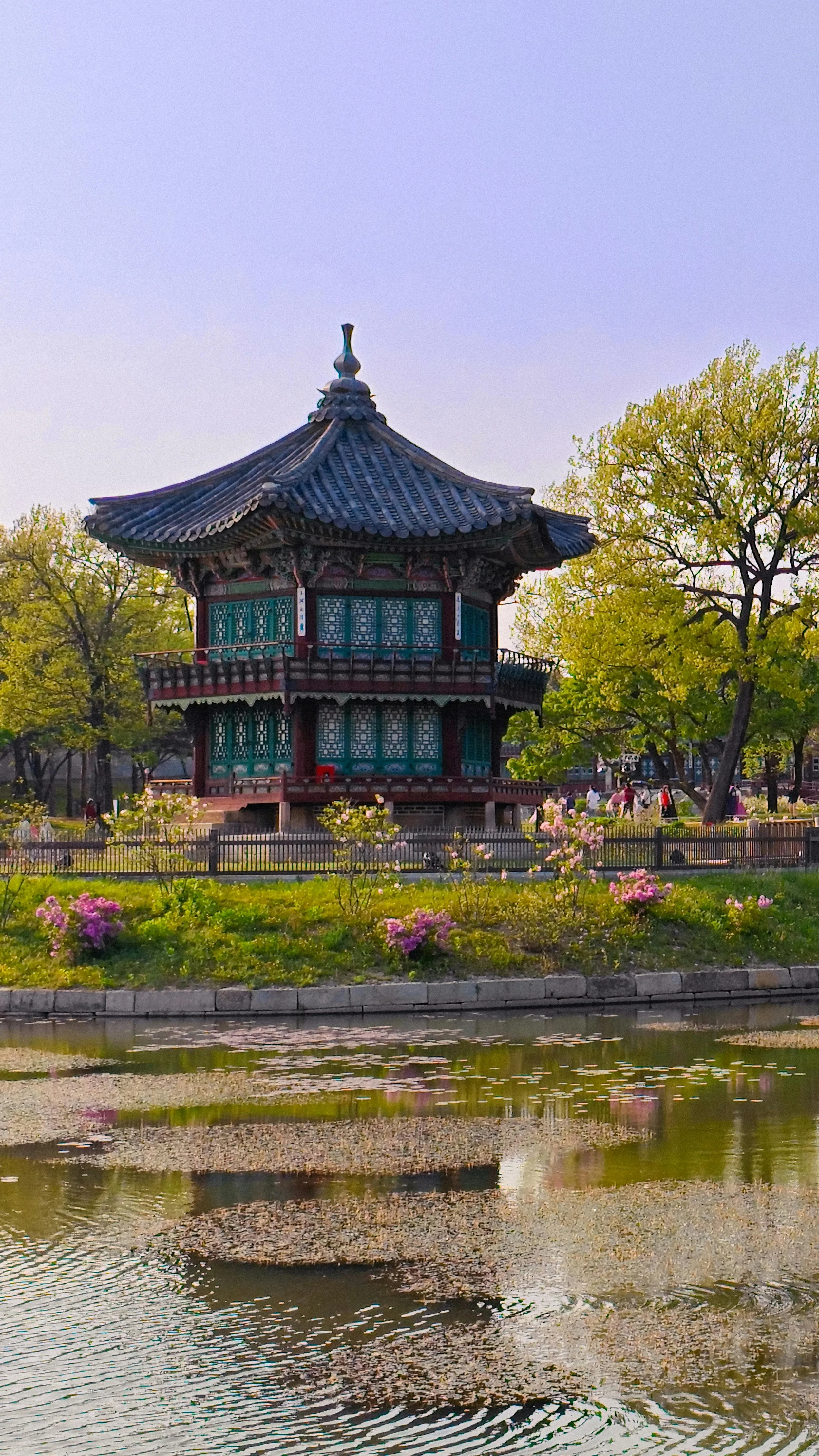 Traditional Asian-style pavilion with curved roof, blue and red wooden details, surrounded by trees and pink flowers, with water in the foreground reflecting the scene.
