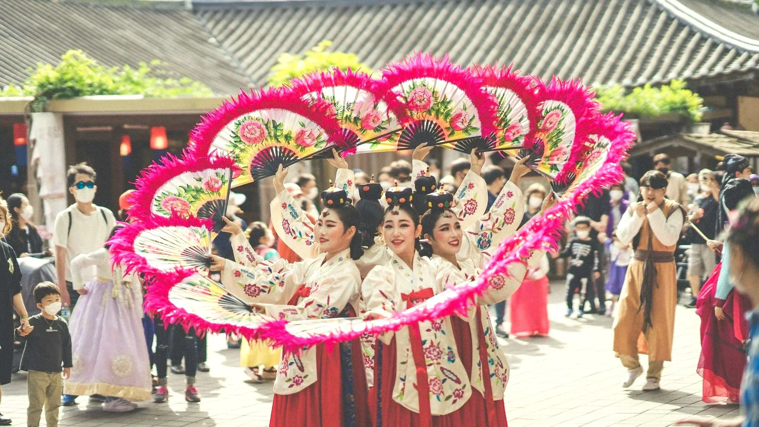 Group of dancers in traditional Korean hanbok performing with colorful fans, surrounded by a crowd in an outdoor setting.