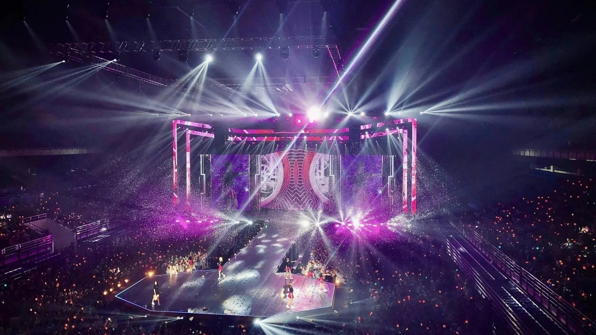 A large indoor concert or event venue with bright colorful stage lights and smoke effects. The stage has a dynamic, geometric backdrop with pink and purple lighting, and there are performers walking on the stage. The audience is visible, with many attendees holding up lighted devices.