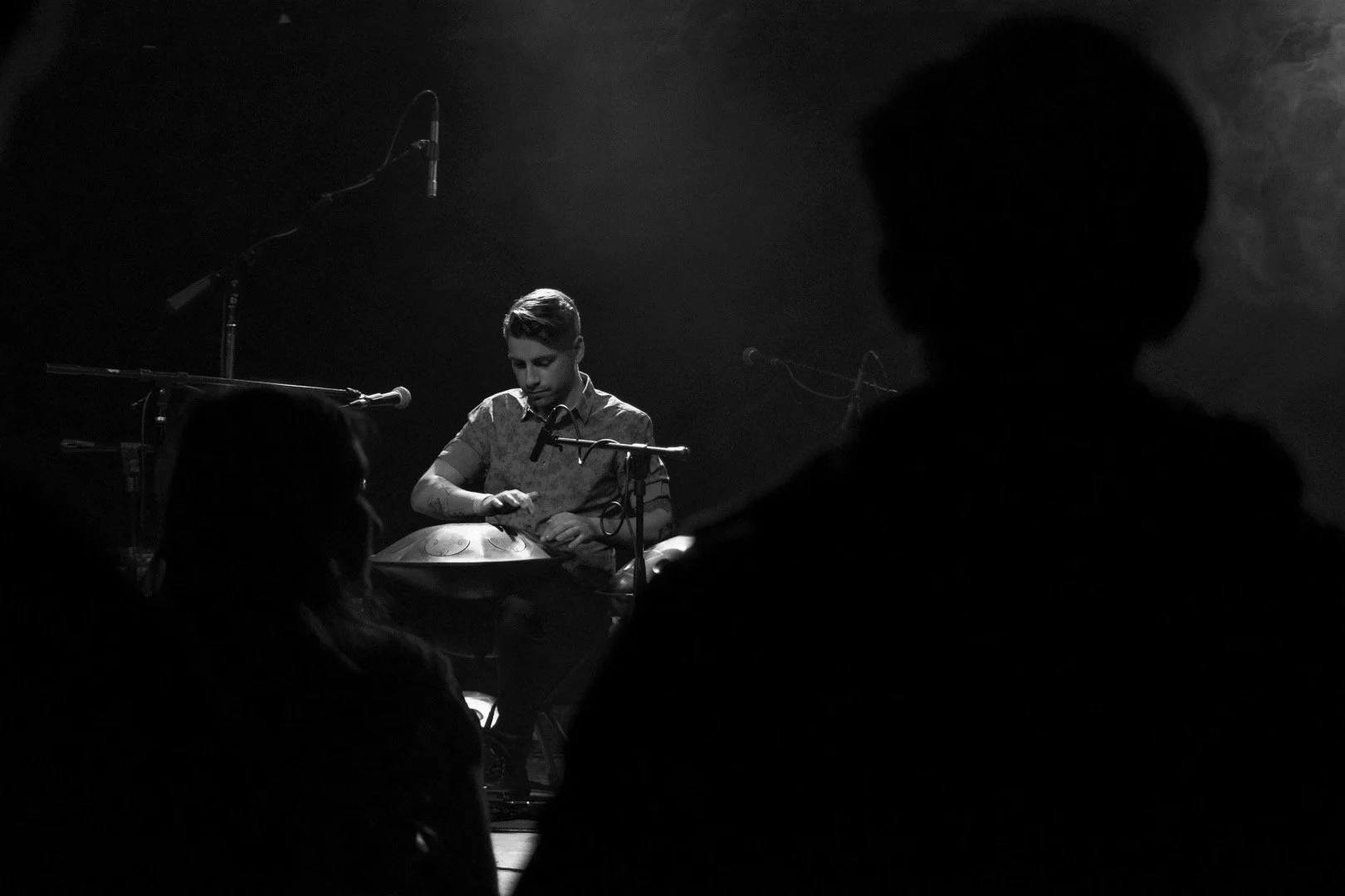 A black and white photo of a musician playing a handpan on stage with several microphones, audience members in the foreground, and stage lighting creating a moody atmosphere.