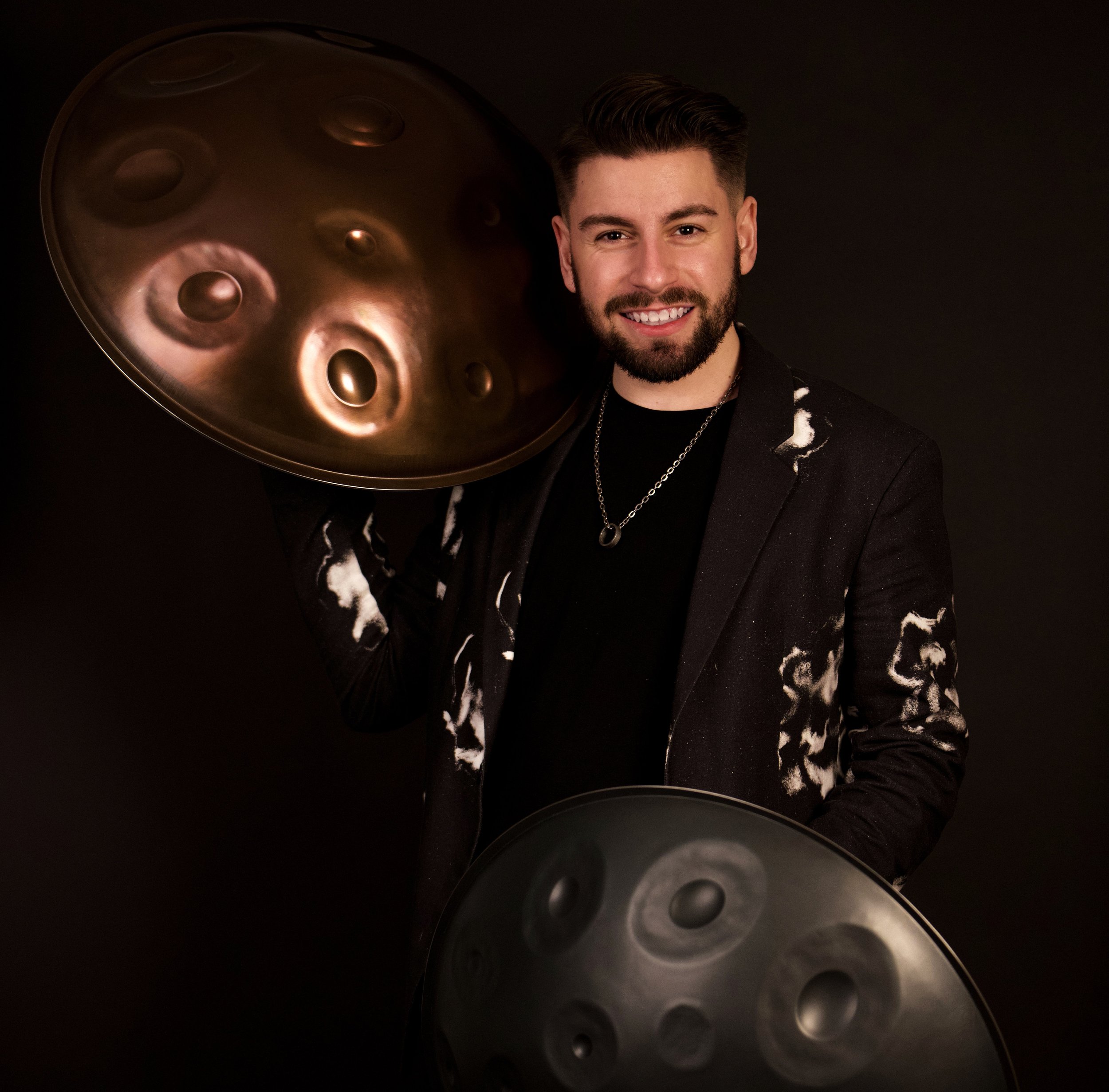 Close-up of a person's face with short, styled brown hair and a beard, smiling, against a dark background, holding a large metallic handpan instrument with circular indentations.