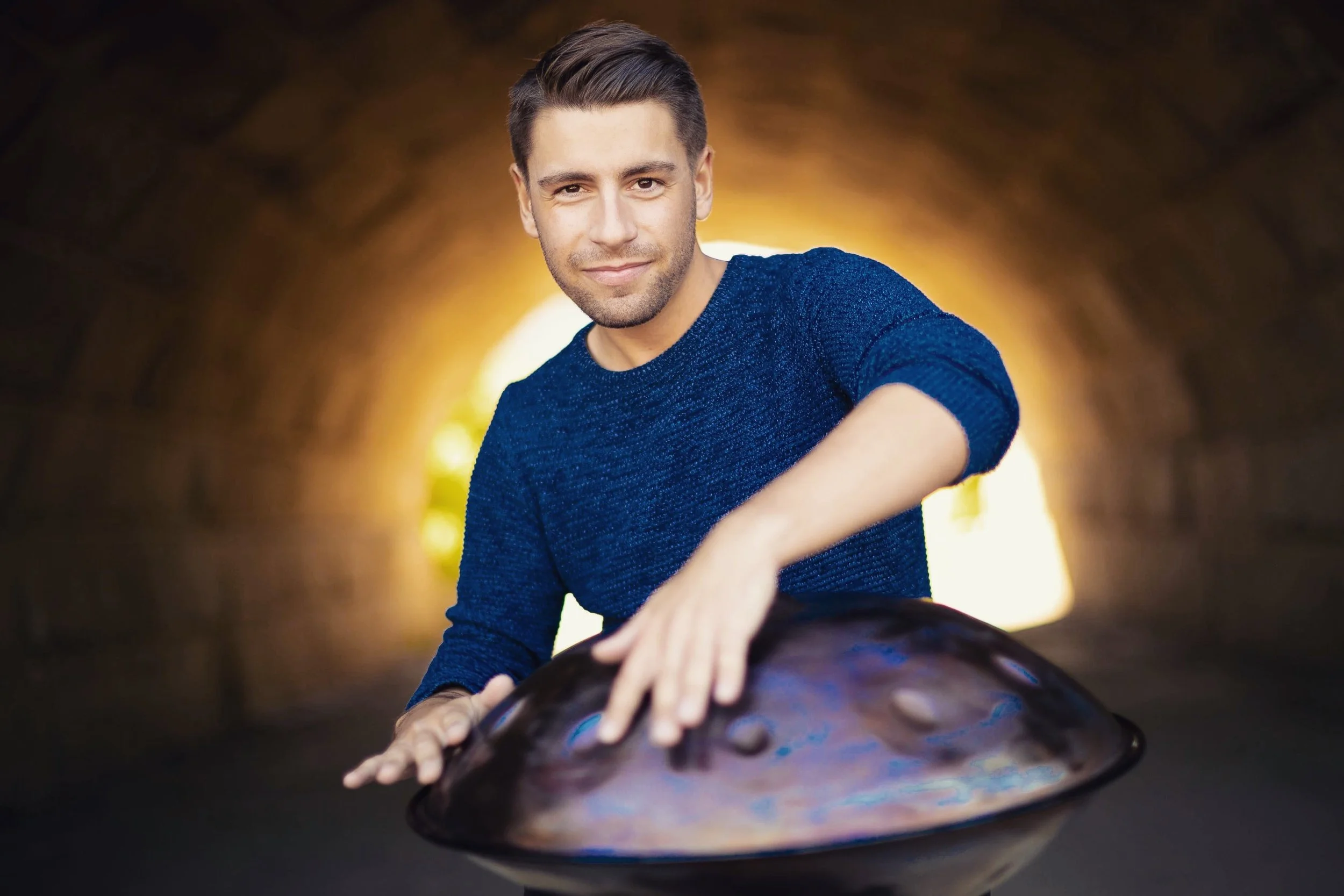 A man with short dark hair and a light beard, wearing a blue sweater, is stretching out his hands on a large, reflective, circular surface inside a tunnel or underground passage with warm, glowing light in the background.