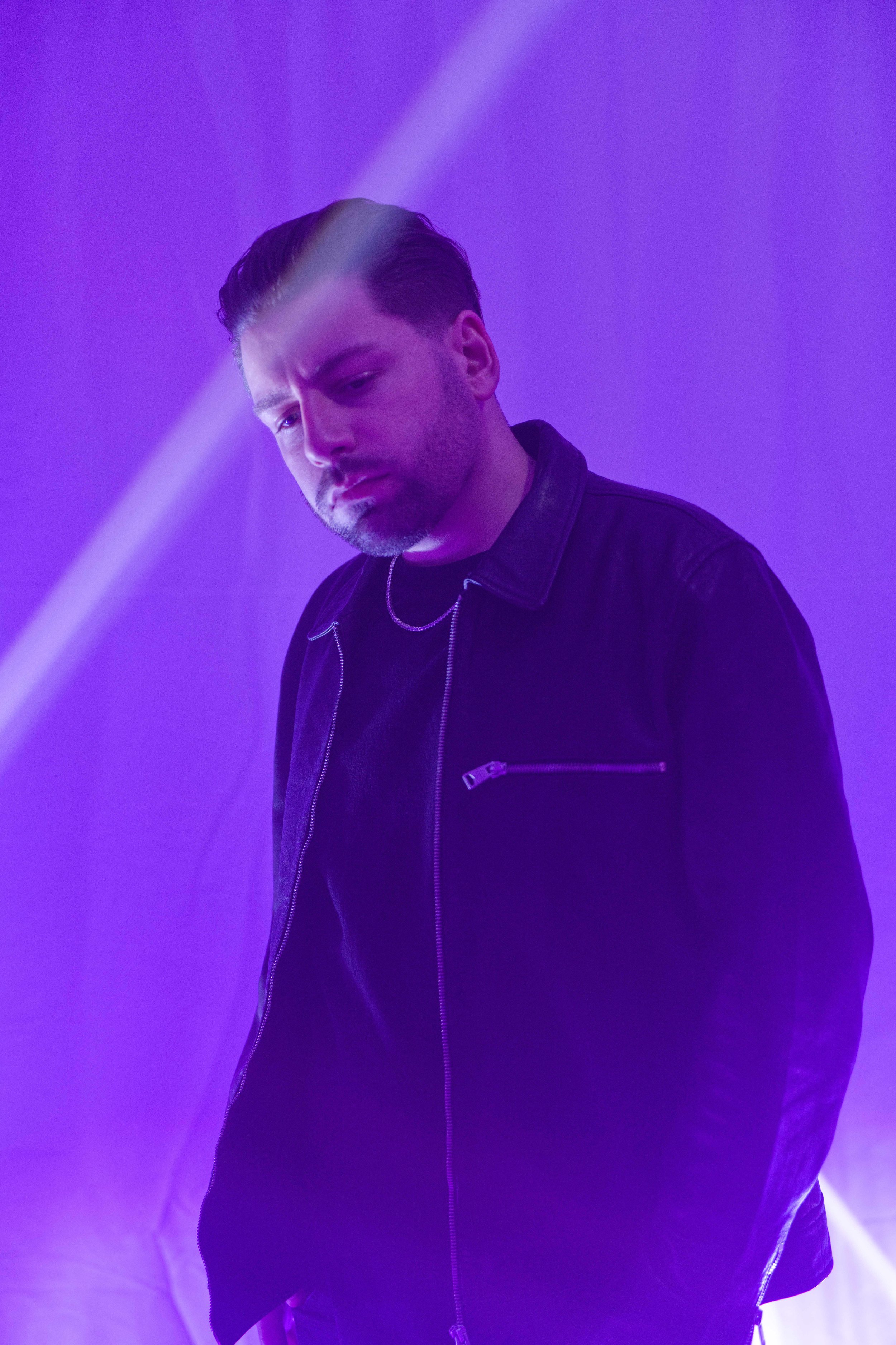 A man with slicked-back hair and a beard in a black leather jacket and chain necklace, looking down, illuminated by purple and blue lighting.