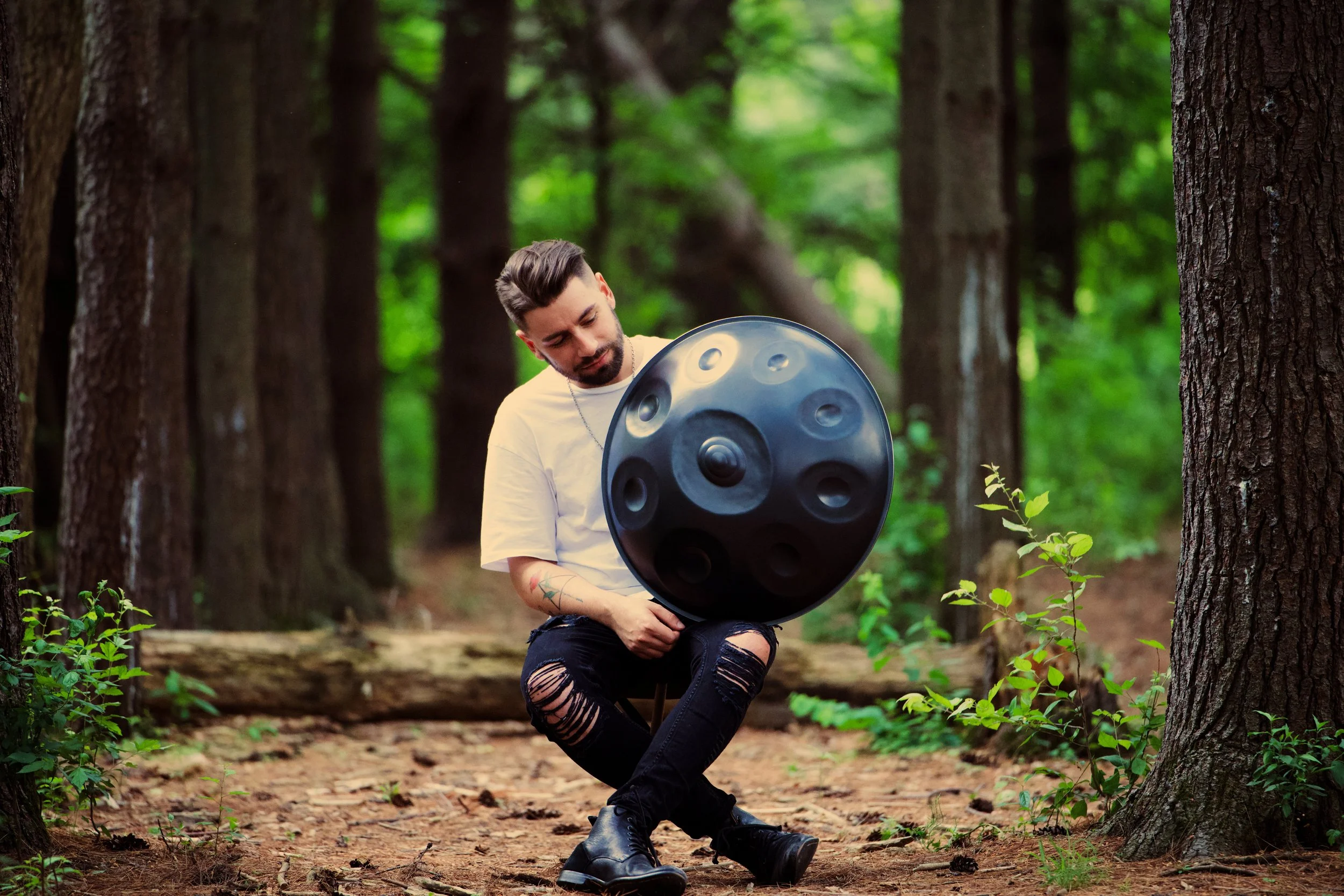 A man sitting in a forest, holding a black steel tongue drum, wearing a white t-shirt, ripped black jeans, black sneakers, with tattoos on his arms, surrounded by tall trees and greenery.