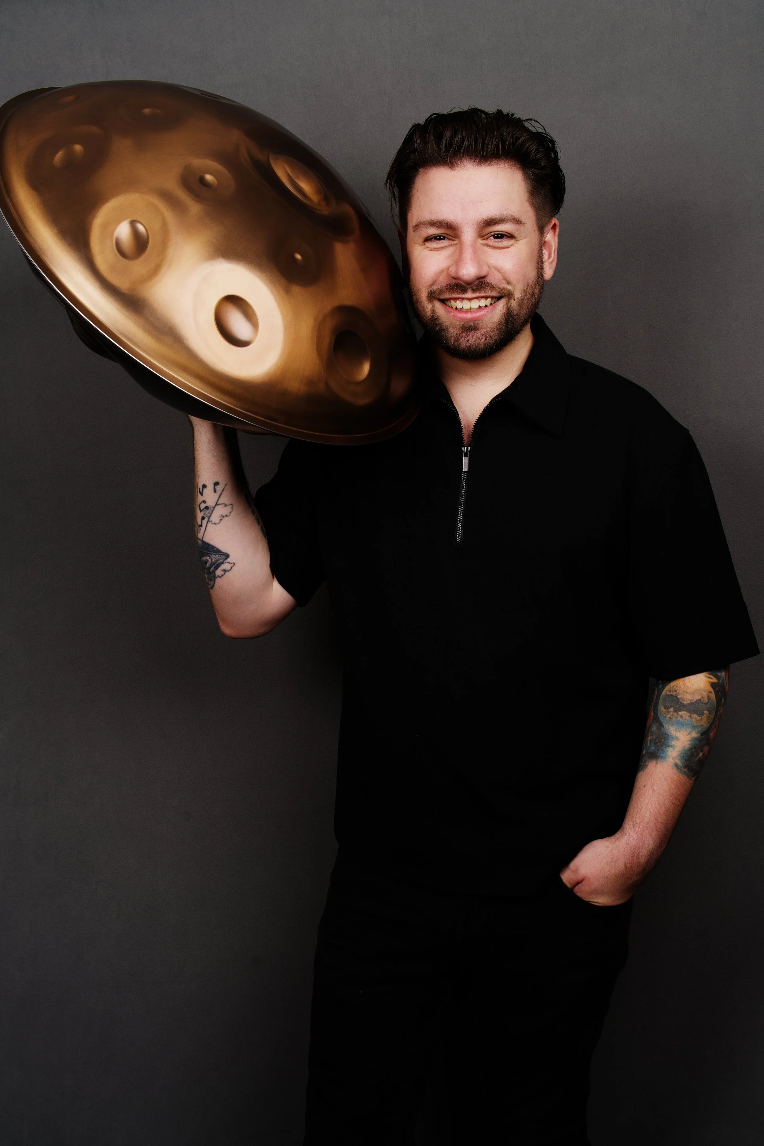 A smiling man with tattoos on his left arm, wearing a black shirt, holding a gold-colored steel tongue drum against a dark gray background.