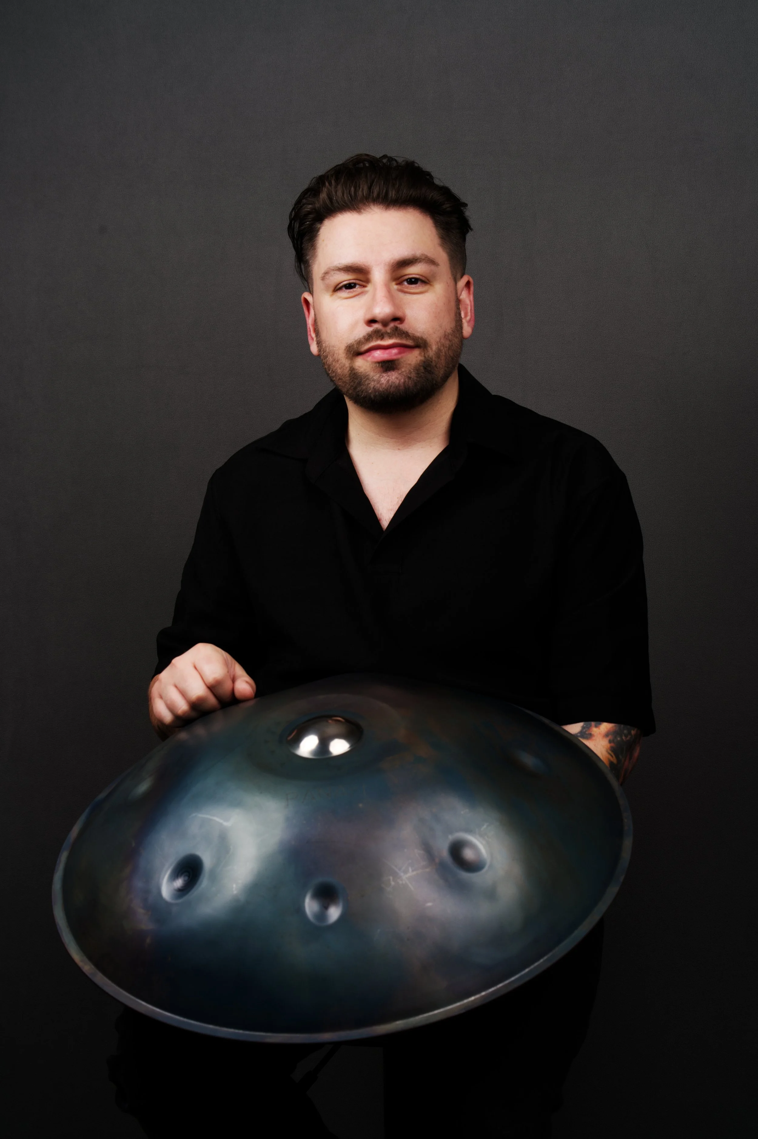 A man with dark hair, a beard, and tattoos on his arm, wearing a black shirt, holding a metallic handpan drum in front of a dark background.