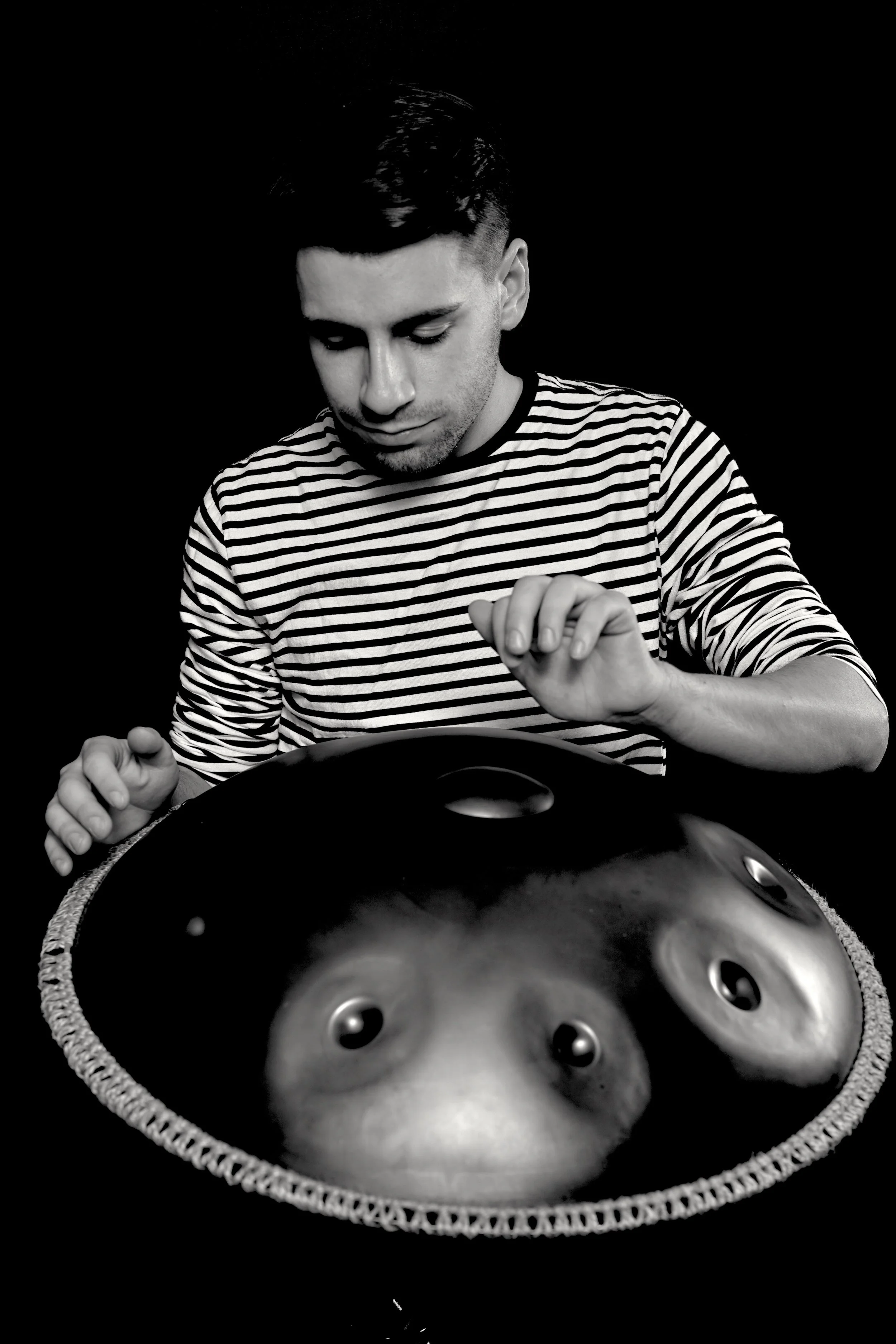 Black and white photo of a man with dark hair and a striped shirt playing a handpan drum, with the drum's reflective surface showing his face.
