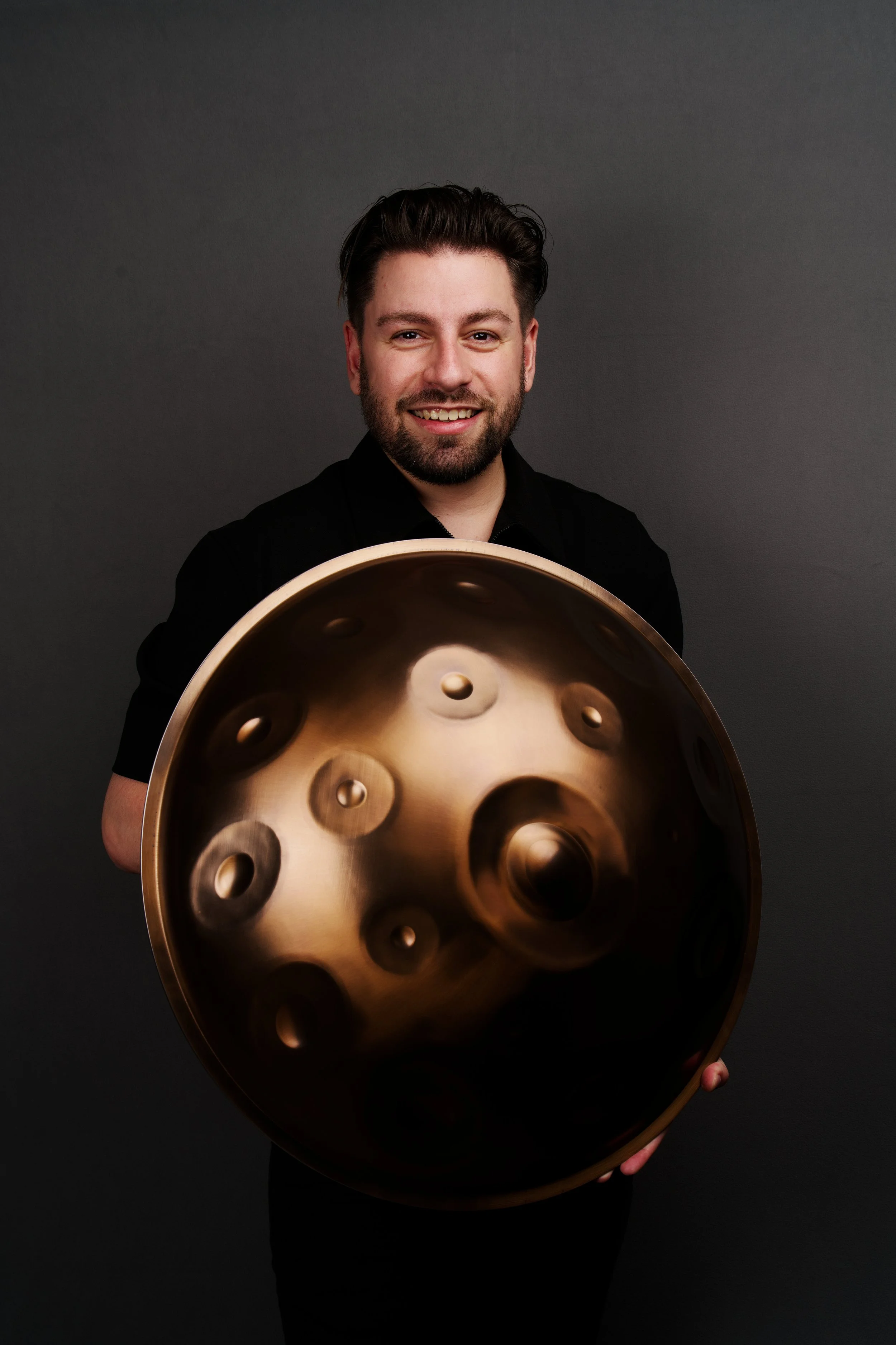 A man with dark hair, beard, and black shirt holding a large bronze handpan musical instrument against a dark gray background.