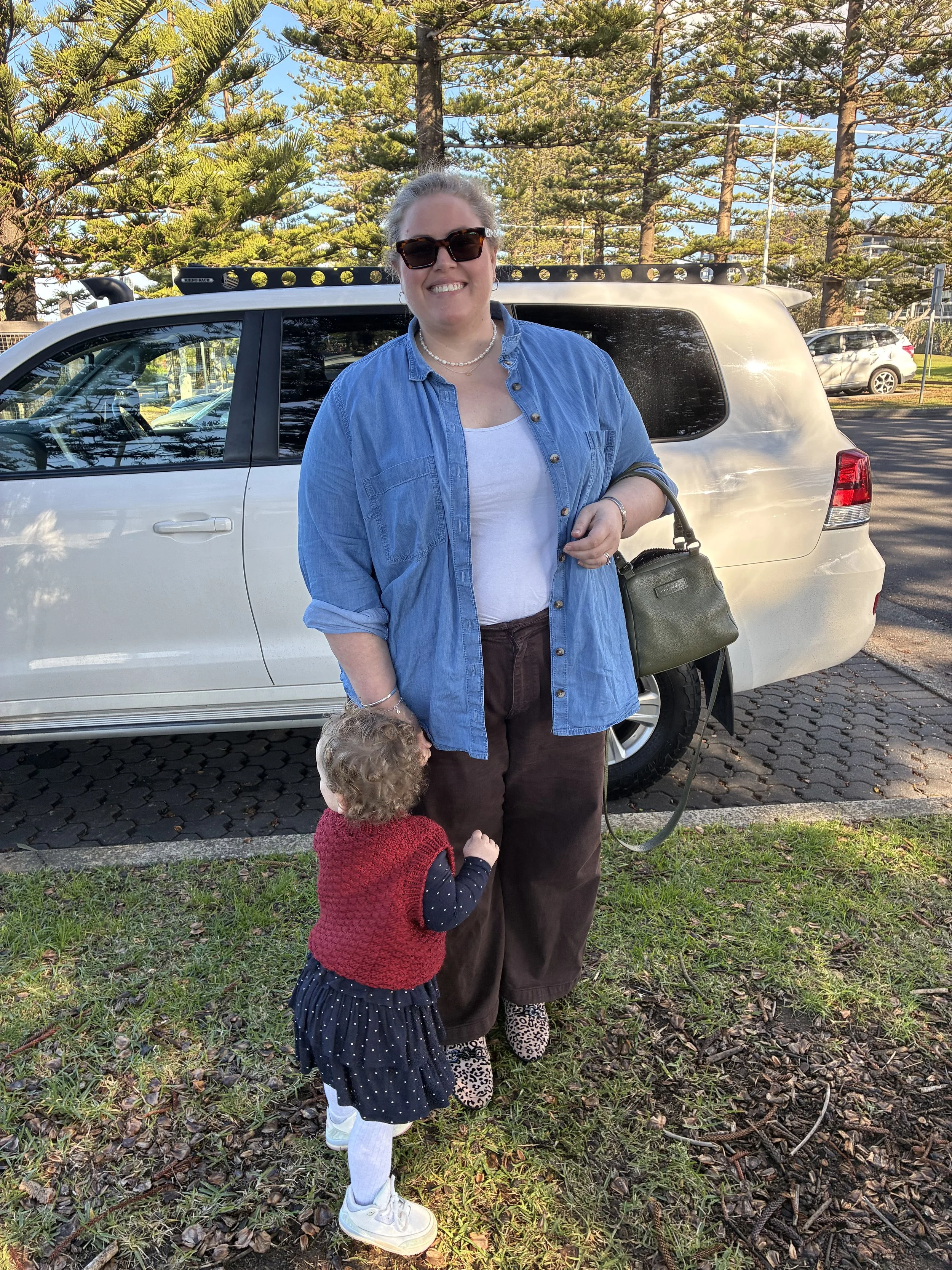 A smiling woman wearing sunglasses, a denim shirt, and jewelry, standing outdoors in front of a white SUV with tall trees in the background. A young girl with curly hair, wearing a red vest and polka dot dress, is reaching up towards the woman, who is holding a handbag.