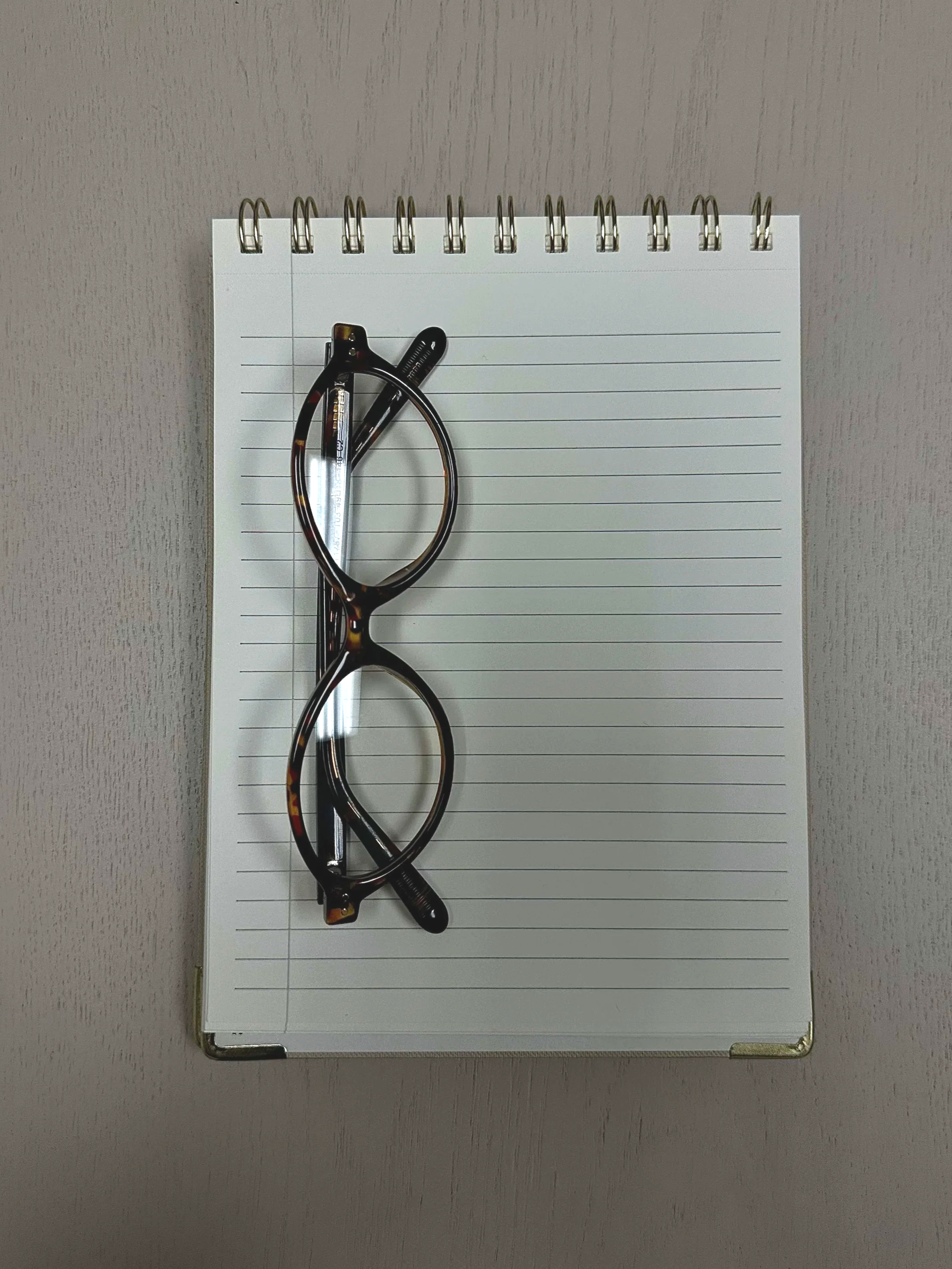 A pair of tortoiseshell eyeglasses, a mechanical pencil, and a black pen resting on a white spiral notepad with lines, placed on a gray surface.