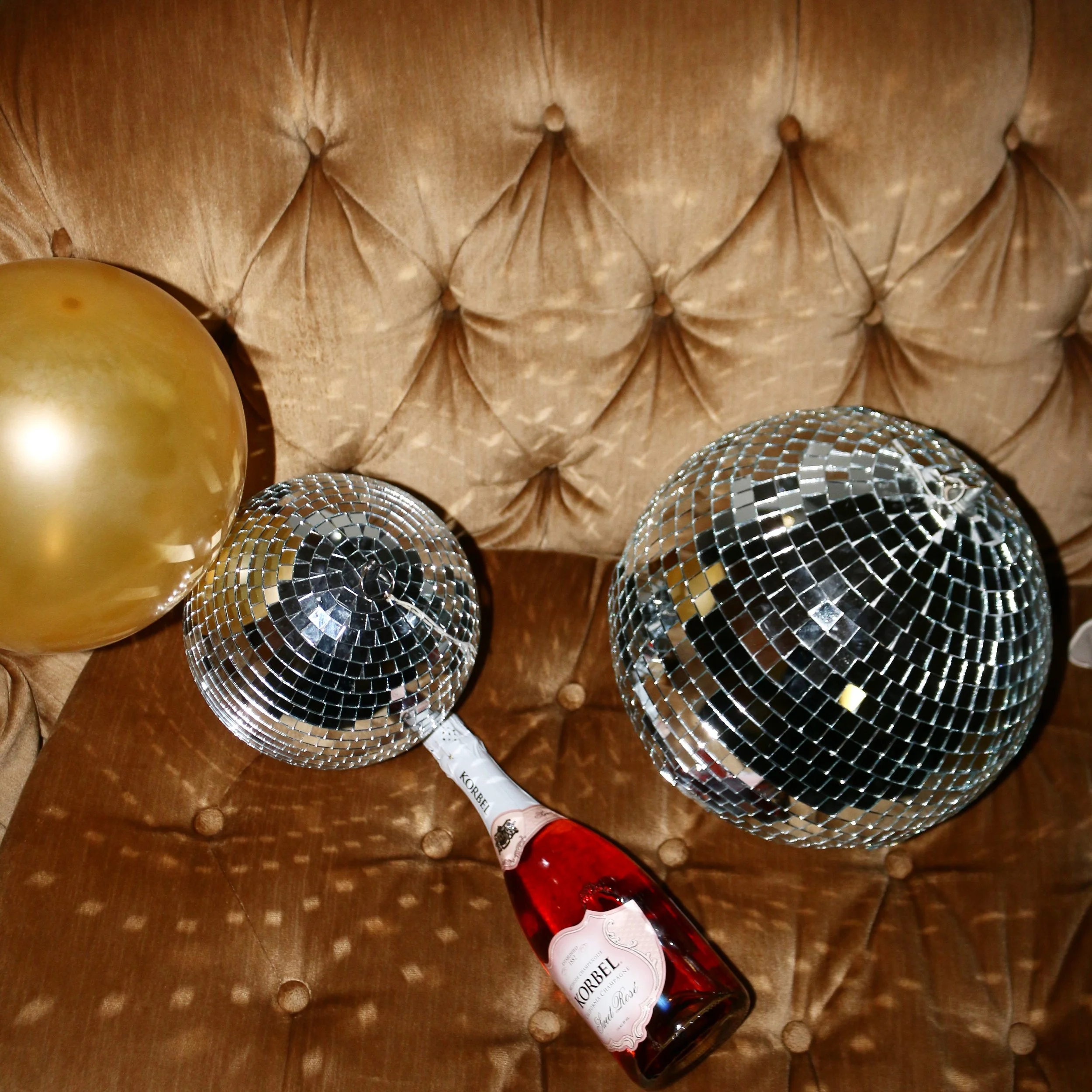 Gold champagne bottle with red foil cap and white label, surrounded by gold and silver Christmas ornaments, on a wooden surface.