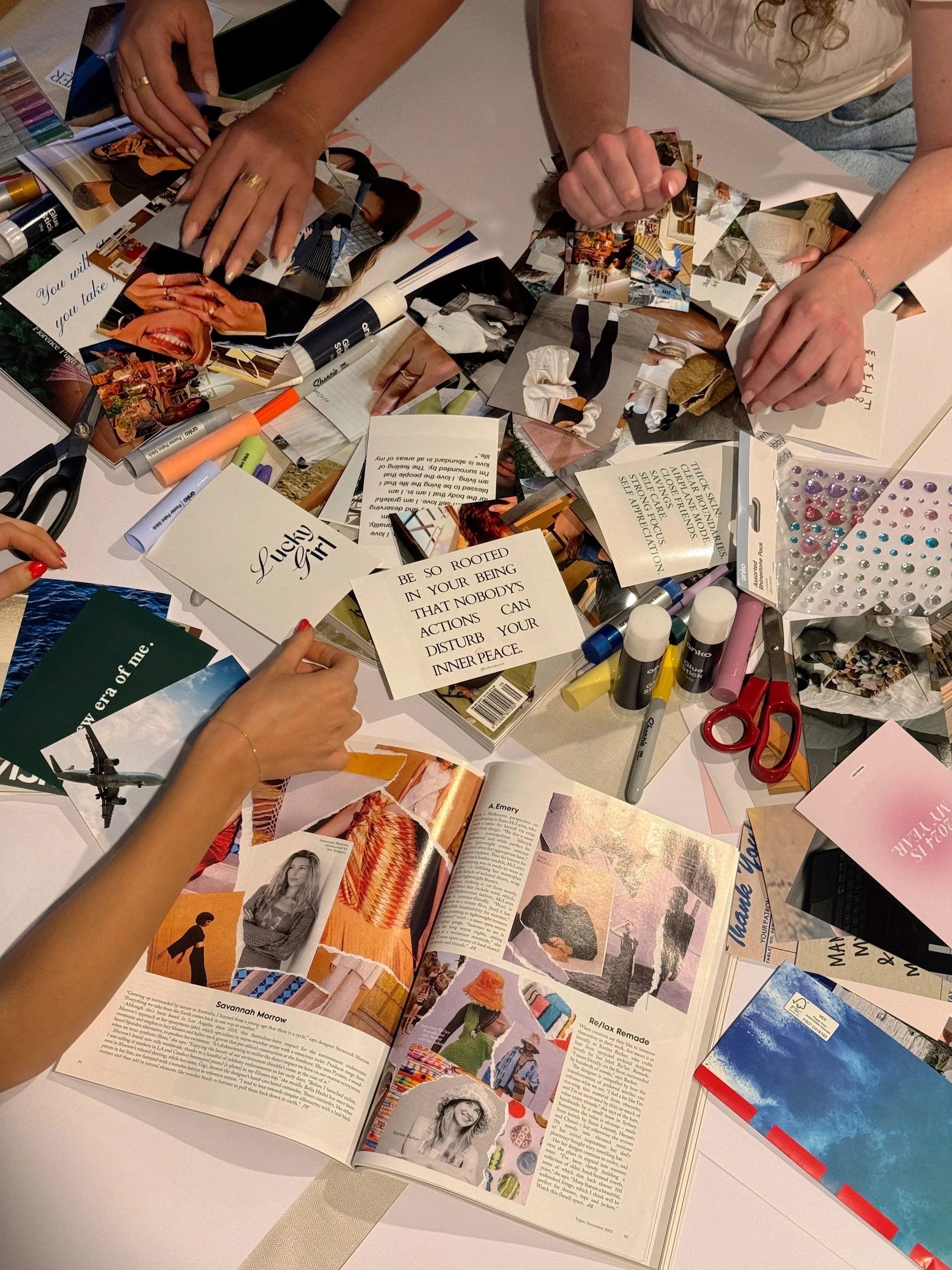 A table covered with magazines, photographs, scissors, glue sticks, and decorative rhinestones, with people arranging images and crafting.