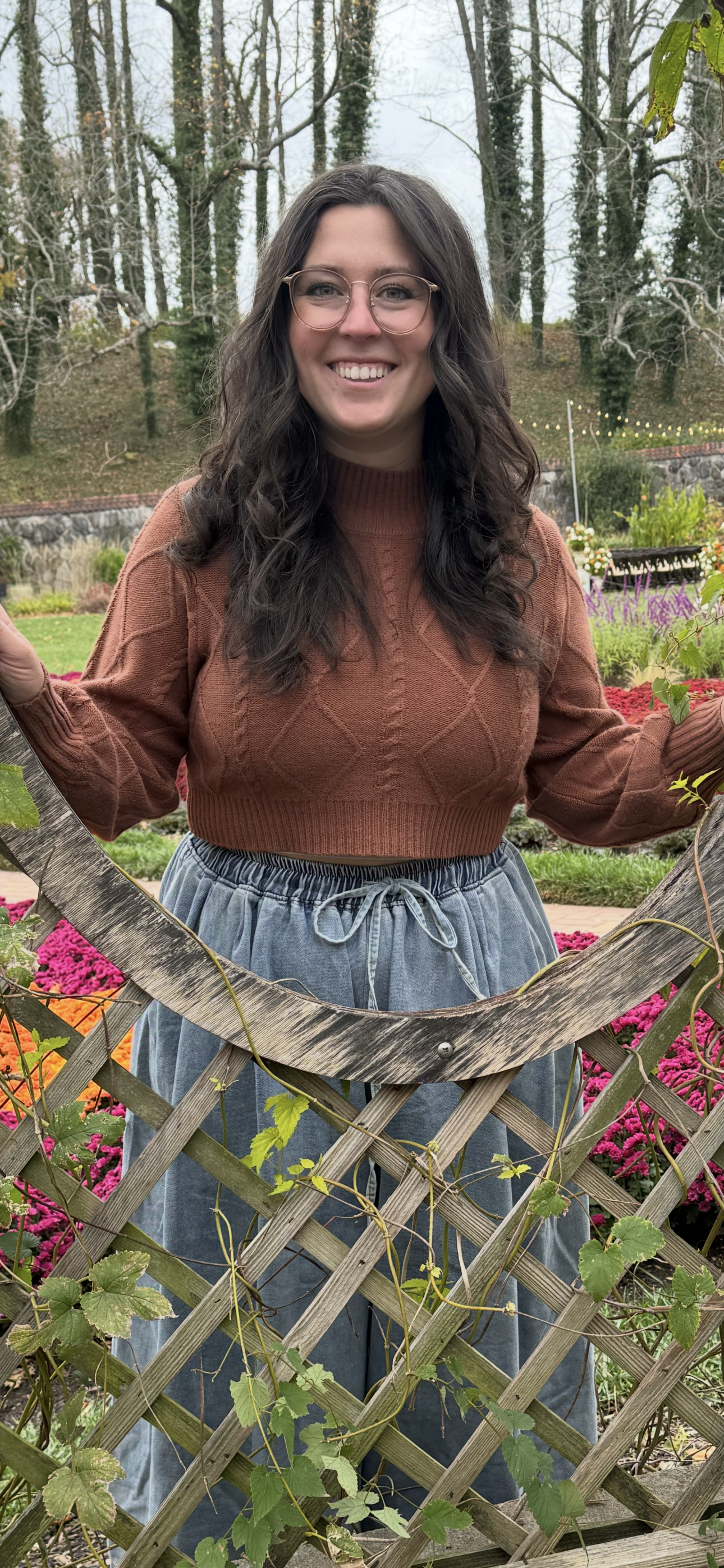 Young woman with glasses smiling outdoors in a garden, standing behind a wooden trellis with green leaves and colorful flowers.