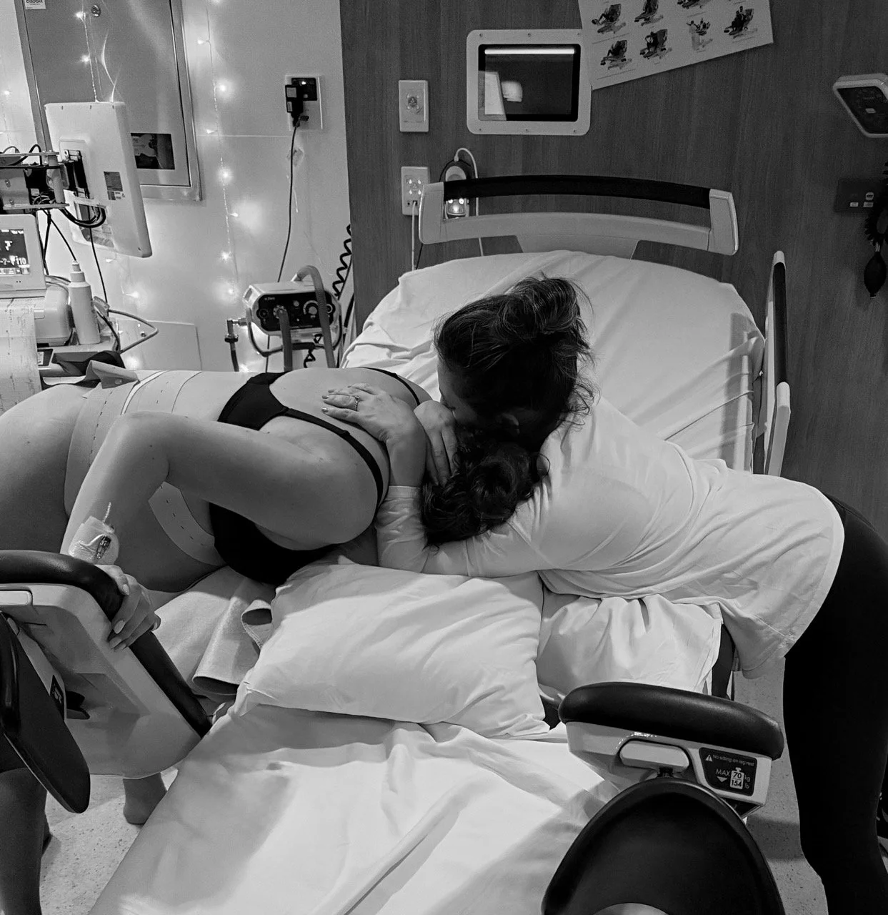 A woman is lying in a hospital bed, being embraced and kissed on the forehead by a person, possibly a nurse or medical professional, in a hospital room with medical equipment and monitors.