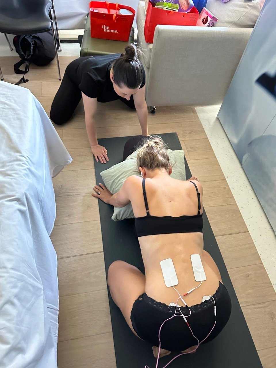 A woman kneeling face down on yoga mat, with her tens machine attached to her back. Steph, is kneeling down next to her, guiding her through labour in their hospital room, or birthing suite setting.