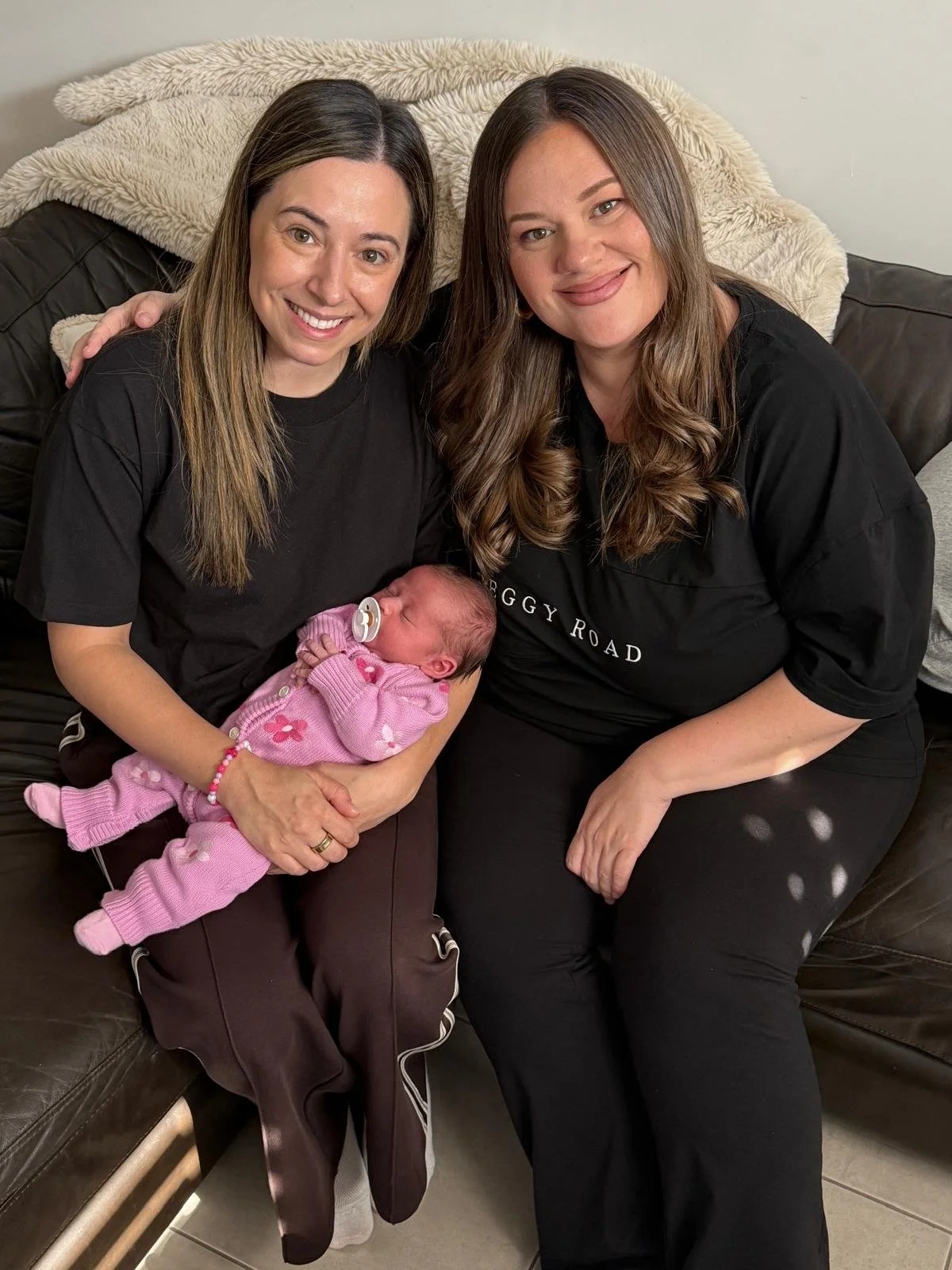 Two women sitting on a dark brown leather couch, one holding a newborn baby dressed in pink. The woman on the left has long brown hair, a smile, and is wearing a black T-shirt and brown pants. The woman on the right has long brown hair, a smile, and is wearing a black T-shirt with white text. They are in a cozy room with a cream blanket on the couch.